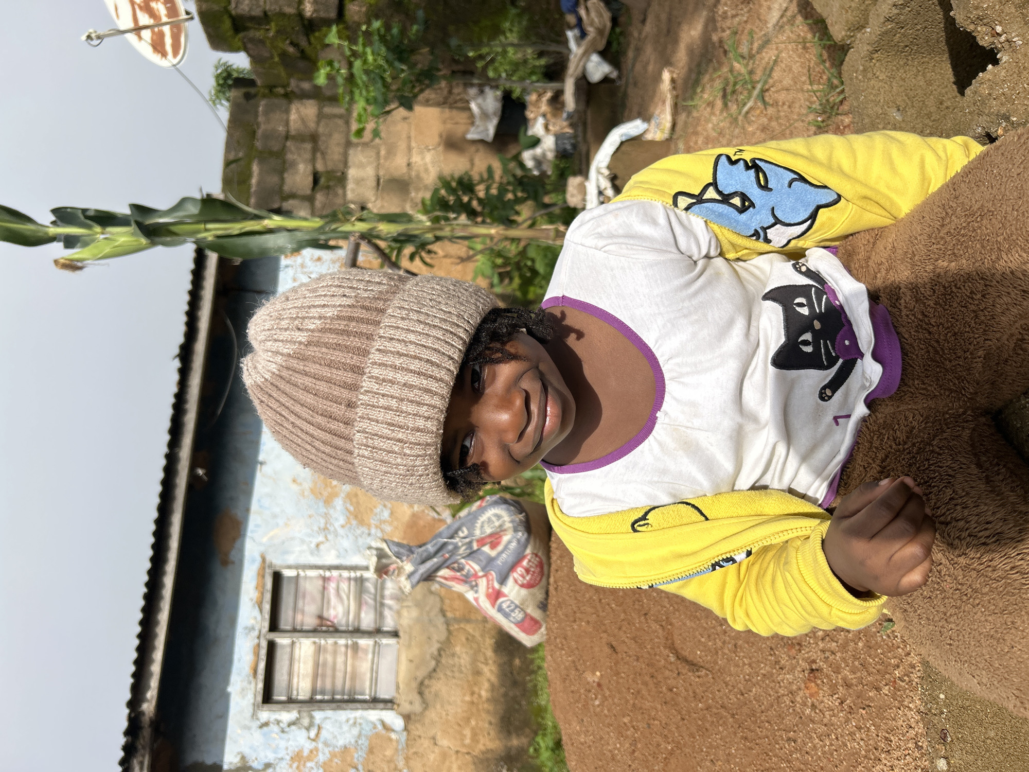 A young Black girl wearing a light brown knit hat, a white t-shirt with a black cat graphic, and a bright yellow jacket with a blue cartoon bird graphic on the sleeve sits in a dirt area in front of a dilapidated building. The building has peeling blue and brown paint and a corrugated metal roof. Behind the girl, a large bag with writing on it and a corn stalk are visible. The ground is covered in reddish-brown dirt. The scene appears to be outdoors, with a cloudy sky in the background. The location is identified as the city of Jos, Nigeria.