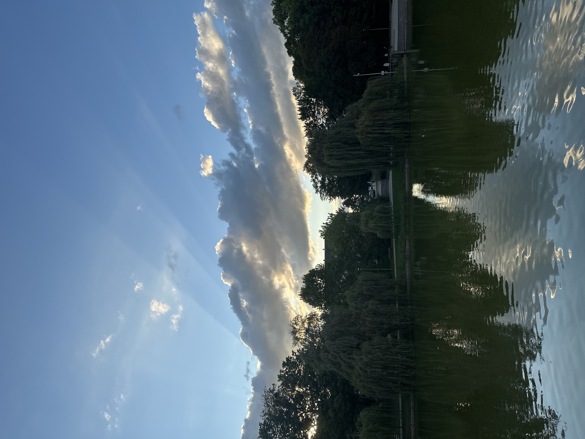 The image shows a body of water, likely a pond or lake, with reflections of trees and the sky. The water is dark green and appears calm, with ripples on the surface. Along the edge of the water, there are several large trees, including weeping willows, with dense foliage. Behind the trees, there is a grassy area and what appears to be a pathway or road with some people walking. The sky above is a gradient of blue and grey, with dramatic sunbeams or crepuscular rays breaking through the clouds. The overall scene suggests a park or natural area within a city, likely during late afternoon or early evening based on the lighting. Without specific landmarks or identifying features, pinpointing the exact location in Racibórz, Poland, is not possible from this image alone, but it depicts a serene outdoor environment.