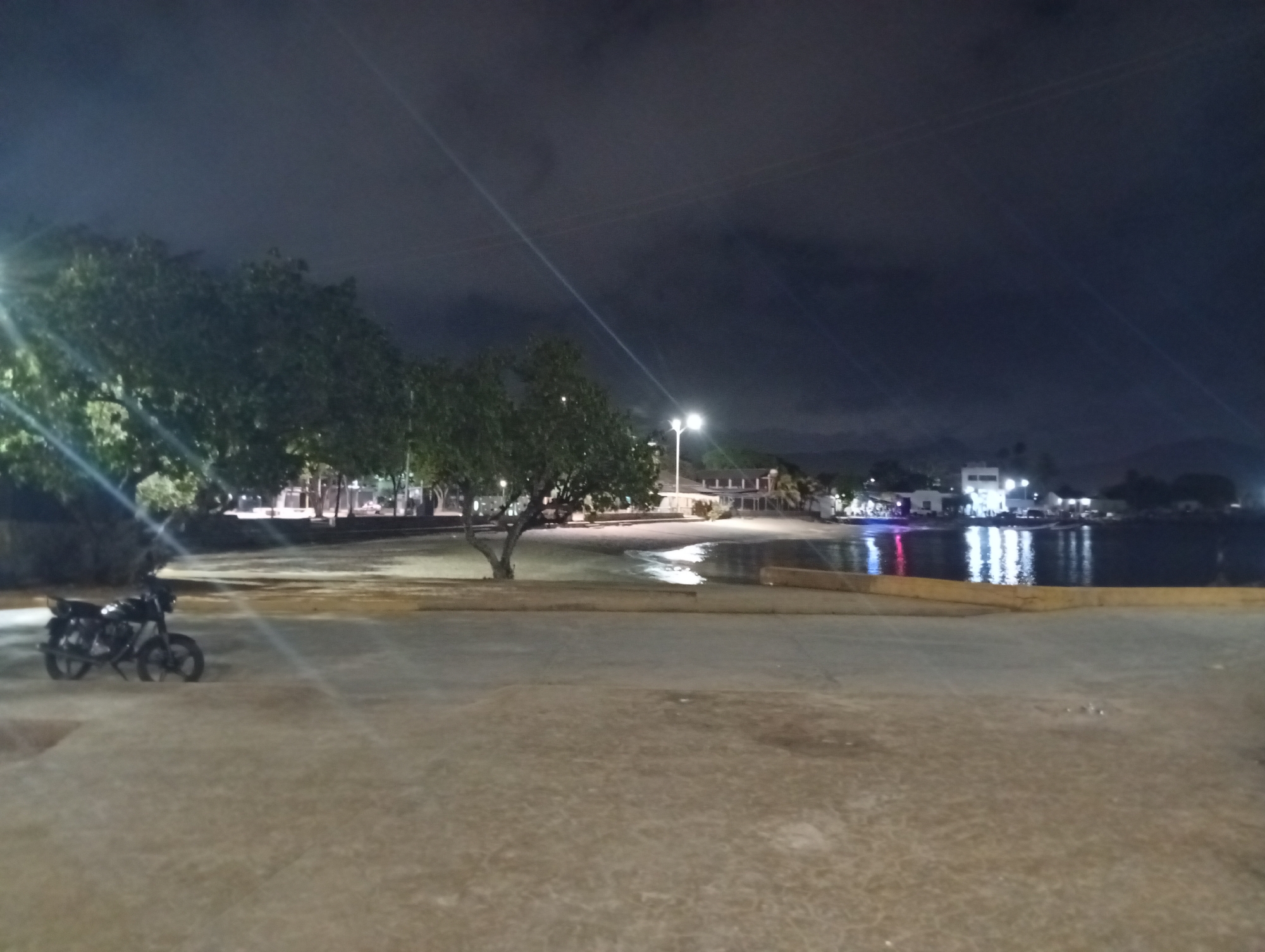 The image depicts a night scene in Juan Griego, Venezuela. In the foreground is a paved area, likely a promenade or parking lot. To the left, two motorcycles are parked near trees. Beyond the paved area, there is a beach and a body of water, possibly the bay of Juan Griego. Reflections of lights can be seen in the water. In the background, there are buildings and lights from what appears to be the town of Juan Griego. The sky is dark with clouds.