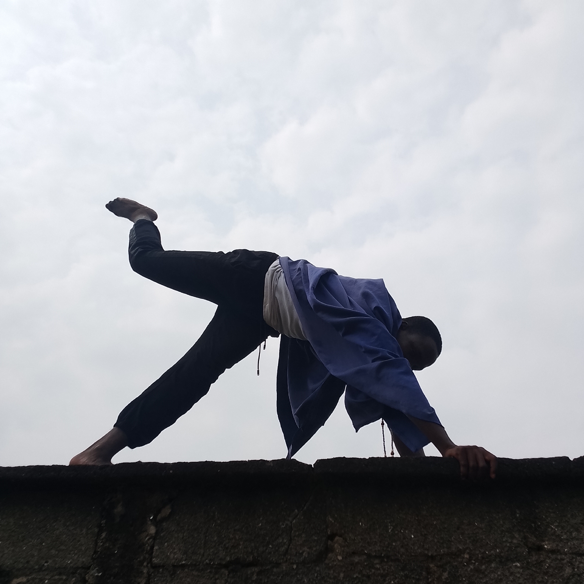 A person is doing a yoga pose on top of a brick wall, with a cloudy sky in the background. The person is wearing dark pants and a blue jacket over a light-colored shirt. Their left leg is extended upwards, while their right leg is bent, and they are balancing on their hands. The scene appears to be outdoors in a city.