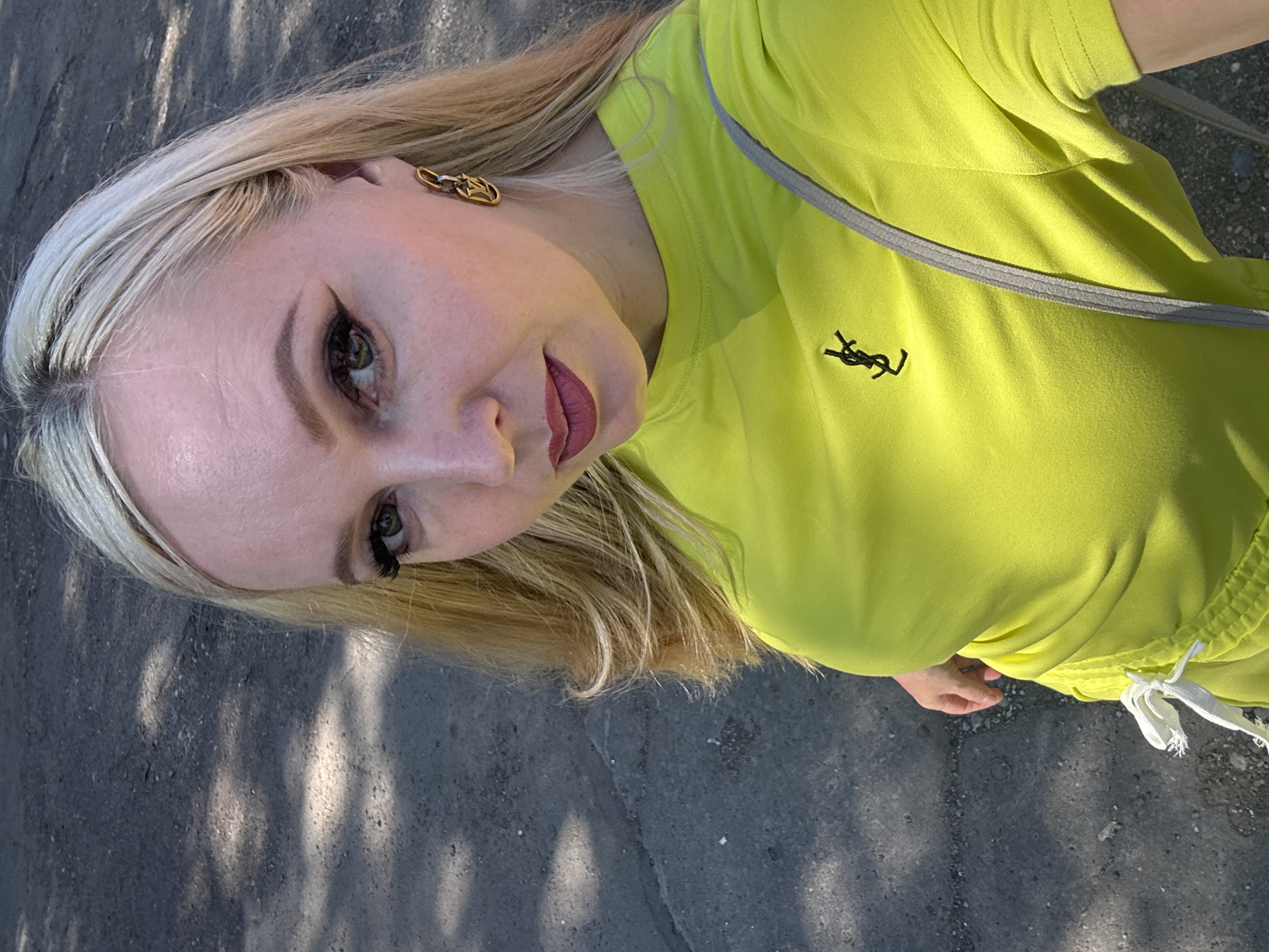 The image shows a woman with blonde hair and light skin wearing a bright yellow t-shirt with a black logo on it. She is wearing gold earrings and has dark eyeliner and lipstick. The background appears to be an outdoor urban setting, possibly a street or sidewalk, with asphalt and dappled sunlight. The woman is looking at the camera and appears to be taking a selfie. No specific location within Komsomolsk-on-Amur, Russia, can be identified from the image.