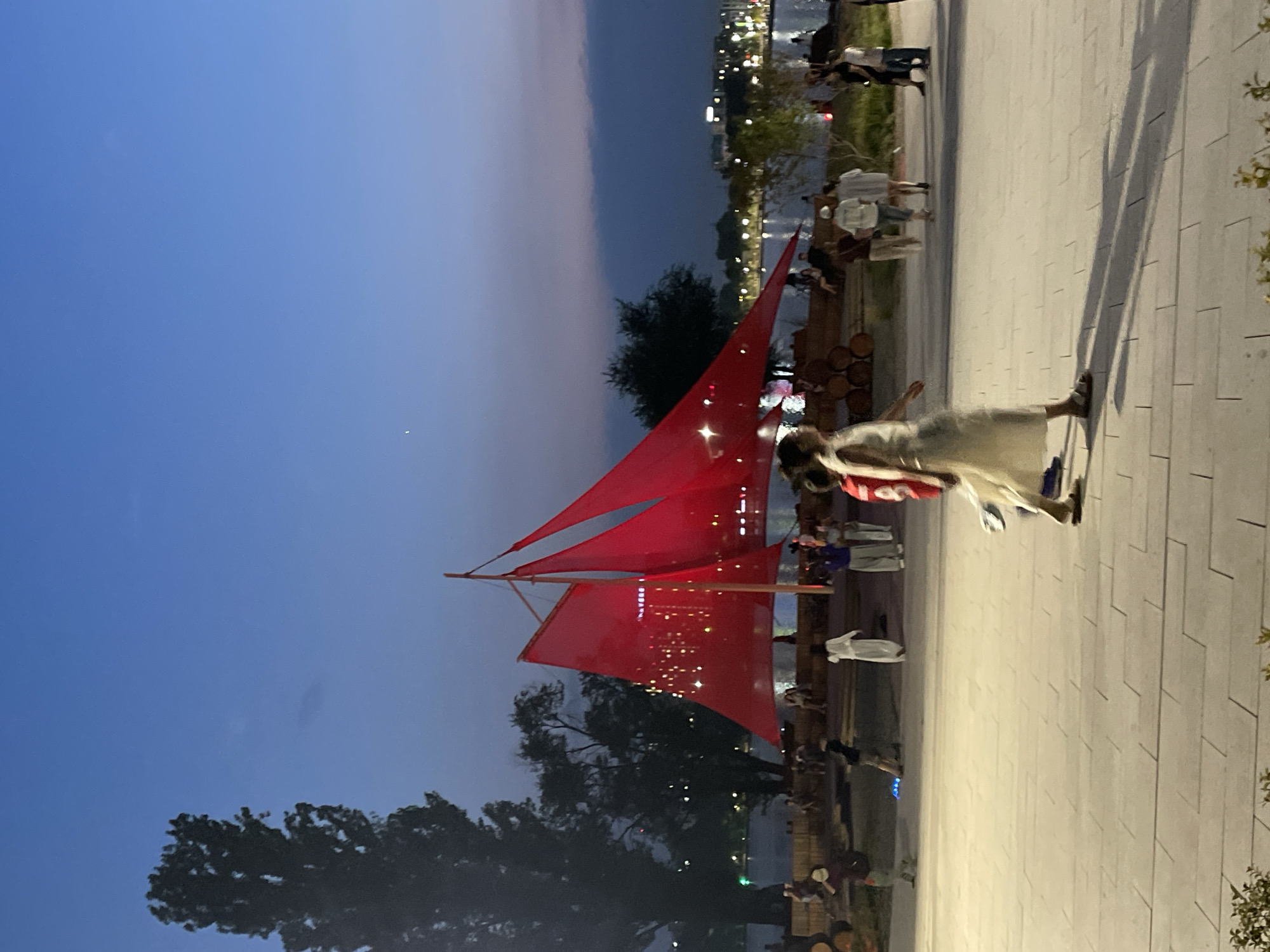 The image depicts an outdoor evening scene in Almaty, Kazakhstan. A prominent, large, red, sail-like structure is the central focal point, adorned with small, bright lights. The structure appears to be an art installation or temporary pavilion, possibly evoking a ship's sail. Several people are visible in the foreground and background, engaged in casual activities. In the immediate foreground, a woman in a light-colored dress is walking towards the right side of the frame, her shadow cast on the paved ground. Further back, other individuals are standing and walking, some clustered in small groups. The environment suggests a public space, possibly a park or waterfront promenade, with a body of water and city lights visible in the distance under a twilight sky. There is a tall tree on the left side of the image, and wooden barrels are stacked near the red structure. The overall atmosphere is relaxed and communal, with the lighting indicating it is after sunset.