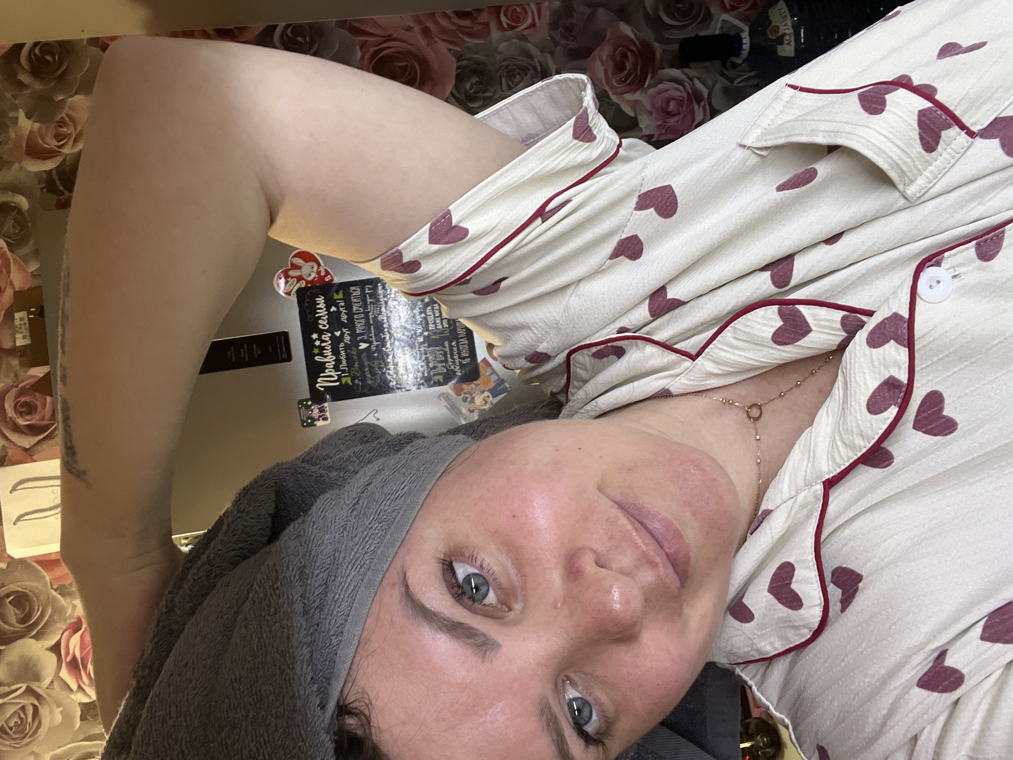 A young woman with blue eyes and light skin is shown in a close-up, selfie-style shot. She is wearing a grey towel turban on her head and a white pajama top with a pattern of dark pink hearts and a red trim. Her left arm is raised, and her hand is resting on the back of her head, revealing her armpit. Her right arm is bent at the elbow, and her forearm is visible. She is wearing a delicate gold necklace with a circular pendant. The background consists of a floral wallpaper with large pink and white roses. A refrigerator with stickers and a handwritten note is visible to the left of the woman. The lighting is warm and soft, suggesting an indoor setting, likely a home. There is no specific information that can be used to ascertain the location within the city of Noviy, Russia.