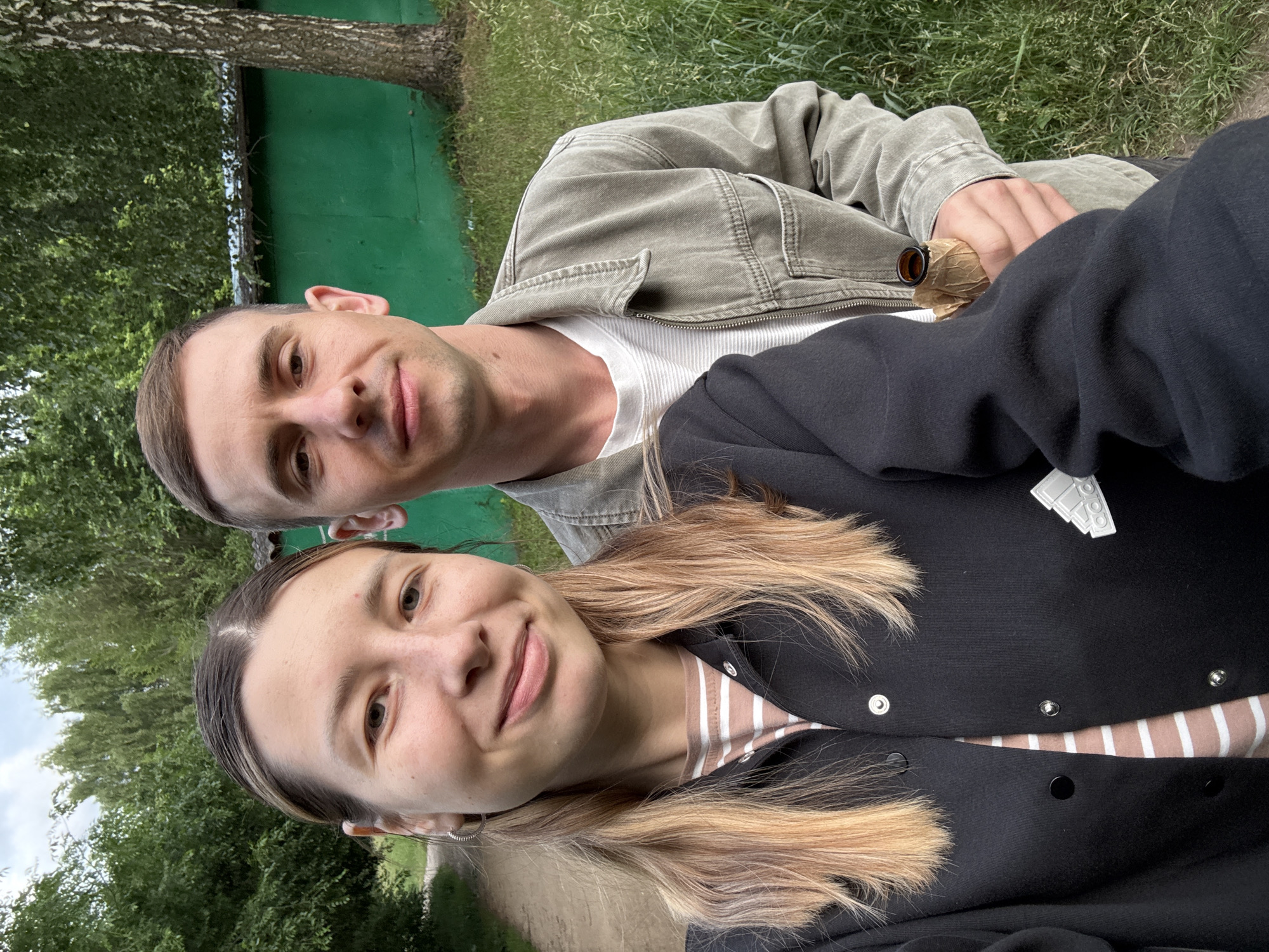 A young couple is taking a selfie outdoors. The woman, on the left, has long blonde hair and is wearing a black jacket over a striped shirt. The man, on the right, has short brown hair and is wearing a green jacket over a white t-shirt. He is holding a bottle wrapped in brown paper. They are standing in front of a green wall and trees. The setting appears to be a park or a rural area, potentially on the outskirts of Moscow, Russia, given the presence of trees and a rustic, unkempt environment. The overall atmosphere is casual and relaxed.
