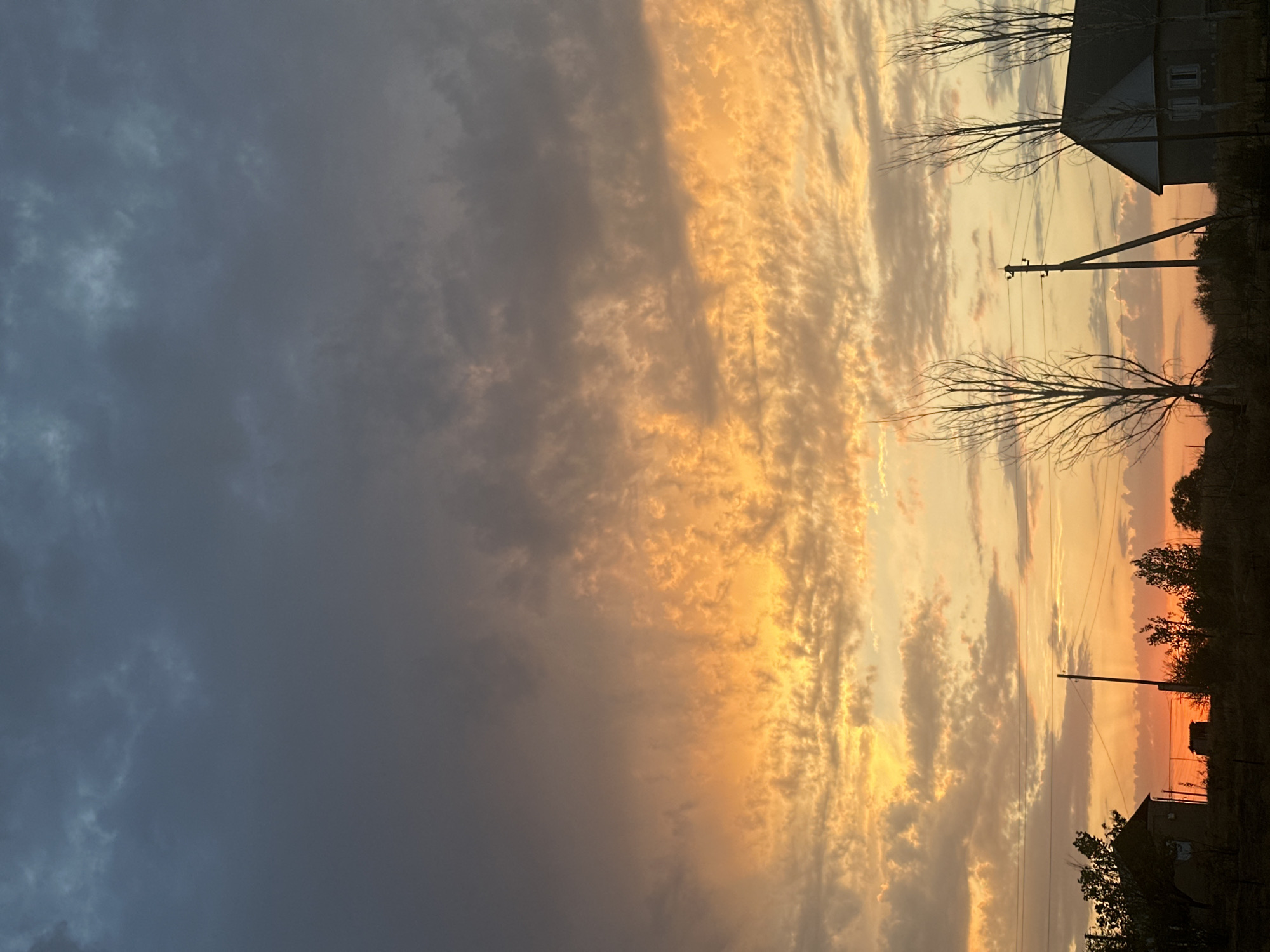 A low-angle, full shot captures a rural landscape under a dramatic twilight sky in Sarymoldayev rural district, Kazakhstan. The sky is filled with a dense layer of clouds, transitioning from a dark, muted gray at the top to vibrant bands of orange and yellow near the horizon. Wispy, textured clouds are illuminated by the setting sun, creating a striking contrast. In the foreground, silhouetted against the bright sky, are several elements of a rural settlement. On the left, a small, dark-colored building with a sloping roof is visible, partially obscured by trees. Further to the right, a utility pole with wires stands prominently. To its right, another building, larger and lighter in color with a dark roof and visible windows, is situated. Between the buildings and the sky, several bare, skeletal trees are silhouetted, their branches stark against the illuminated clouds. The ground is covered with dark vegetation, suggesting a natural or uncultivated area. Power lines are strung across the scene, further emphasizing the rural setting.