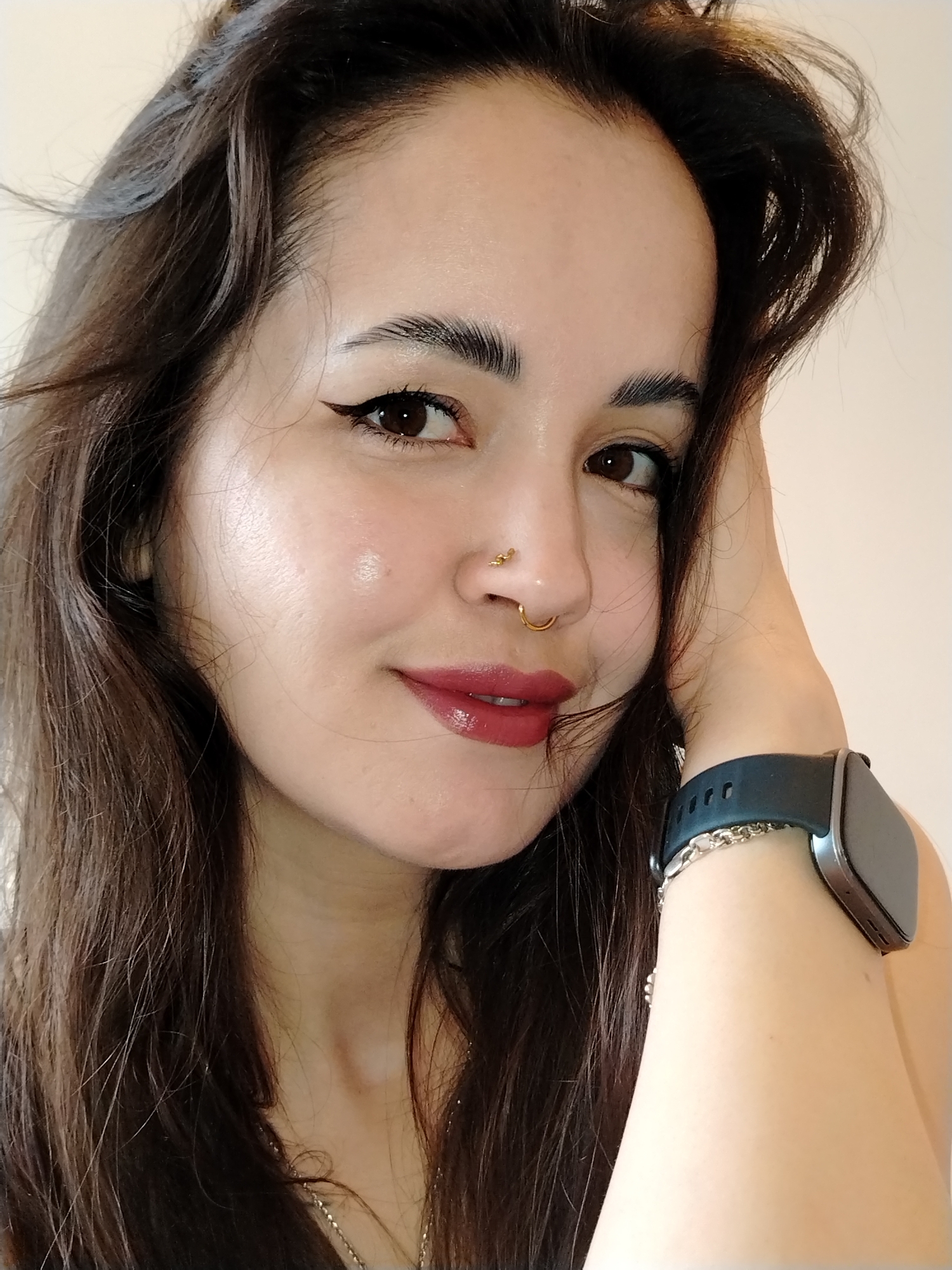 A close-up portrait shows a woman with fair skin and dark brown hair posing with her left hand resting on her head. She has dark eyes with winged eyeliner, thick eyebrows, and a nose adorned with two gold rings in her left nostril. Her lips are painted with a dark red, glossy lipstick. She is wearing a black smartwatch on her left wrist, accompanied by a silver chain bracelet. A delicate silver necklace is partially visible at her neck. The background is a plain, light-colored wall. The location is unknown.
