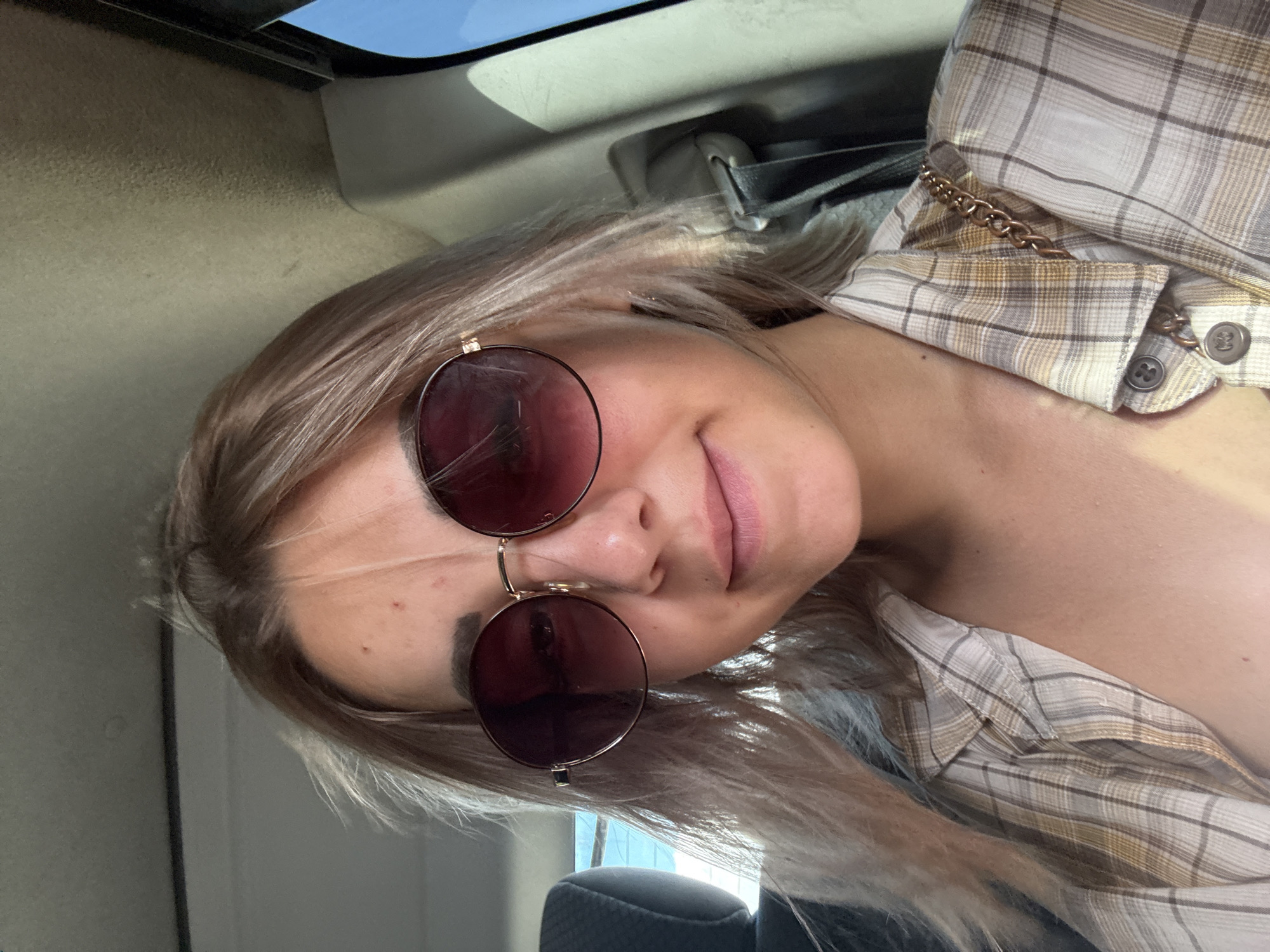In the city of Yuzhno-Sakhalinsk, Russia, a young woman with blonde hair, wearing round sunglasses and a plaid shirt, sits in the passenger seat of a vehicle. The lighting suggests it is daytime, possibly sunny, with light filtering into the car. The woman is looking directly at the camera, with a slight smile. The interior of the car is visible, including the car's headliner and a seatbelt. A portion of the woman's shoulder and neck are also visible, and she is wearing a chain strap accessory over her shirt.