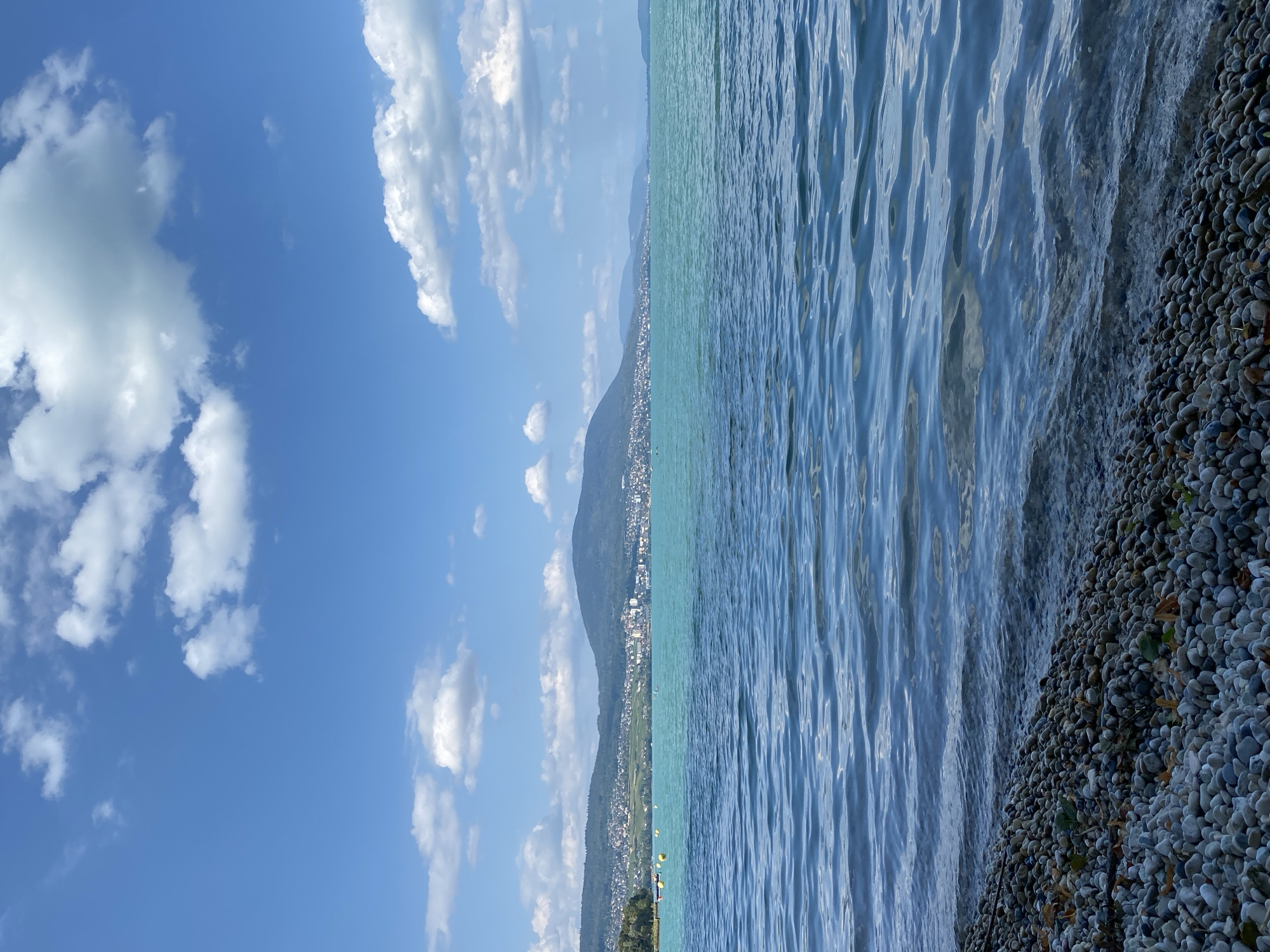 The image depicts a scenic view of Lake Neuchâtel with the town of Cortaillod, Switzerland, visible in the background. In the foreground, a pebble beach lines the shore, meeting the calm, blue waters of the lake. The water exhibits ripples and reflections of the sky, suggesting a gentle breeze. In the mid-ground, the town of Cortaillod is spread along the lakeshore, nestled at the base of rolling green hills that form the backdrop. Buildings of various sizes and colors are discernible within the town, and a few boats are present on the water. The sky above is a vibrant blue, dotted with fluffy white clouds. The overall impression is one of a peaceful, sunlit day in a picturesque lakeside setting.