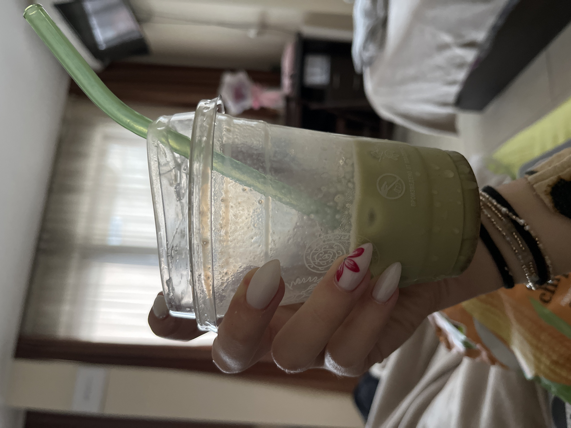 A woman's hand, adorned with manicured nails, holds a plastic cup filled with a green beverage, likely matcha or a similar drink. A green straw is inserted into the cup. The hand has a few bracelets, but the exact number and style aren’t completely discernible. The background suggests an interior setting, potentially a bedroom, with a bed, a TV, and other furniture items. The specific location within Sofia, Bulgaria, cannot be determined from this image.