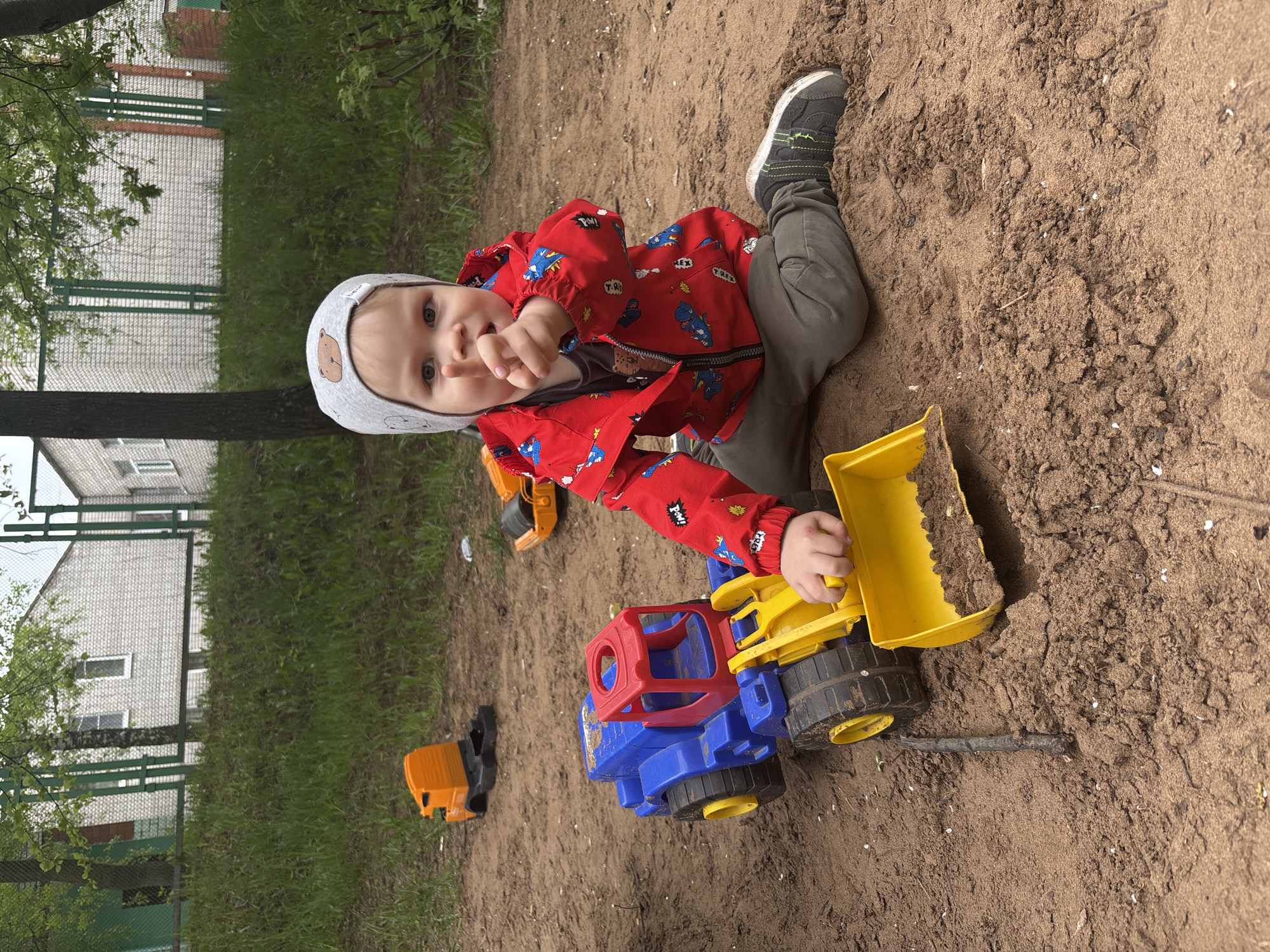 The image shows a young child, possibly around one to two years old, playing in what appears to be a sandbox. The child is wearing a light gray hat with a bear design, a red jacket with cartoon dinosaur or monster images, and gray pants. The child is positioned close to the camera, facing slightly to the left, with one hand extended.
In front of the child is a large, colorful plastic toy truck, primarily blue, yellow, and red. The truck is on the sand, and the child appears to be interacting with it. To the left of the truck, there are at least two other smaller orange or yellow toy vehicles.
The background consists of greenery and what looks like a brick or concrete building. The location seems to be a park or playground area.
Without specific indicators, it is not possible to confirm the exact location, but nothing visible in the image provides information to confirm or deny it being in Izhevsk, Russia.