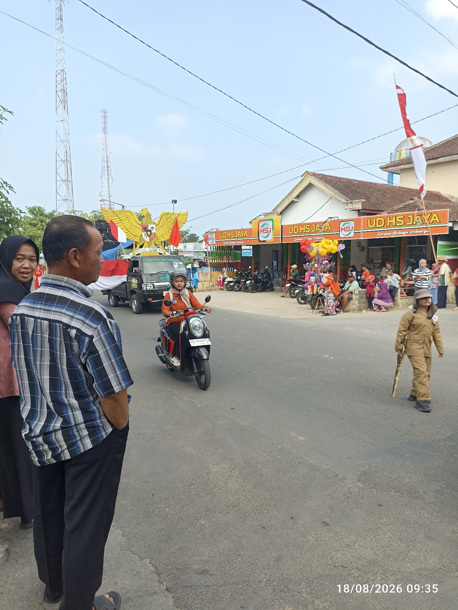 In Munjungan, Indonesia, on August 18, 2026, at 09:35, a street scene unfolds with elements of a parade or celebration. In the foreground, a man in a blue and white checkered shirt and black pants stands with his hands in his pockets, facing away from the camera, while a woman in a black hijab and light pink top stands to his left, looking towards the parade.
The parade features a black pickup truck adorned with a large yellow Garuda Pancasila emblem and Indonesian flags. A woman in an orange jacket and headscarf is riding a black scooter in front of the truck, wearing a helmet. Further back, a child in a tan uniform and hat, carrying a wooden stick resembling a rifle, walks along the road.
On the right side of the street, a building with a sign reading "UD HS JAYA" is visible, with several people gathered outside. Some are seated, while others stand watching the parade. A collection of colorful balloons is also present near the building. Motorcycles are parked along the side of the road. In the background, two tall communication towers rise against a clear blue sky with thin clouds. The road surface is asphalt with visible wear and tear.