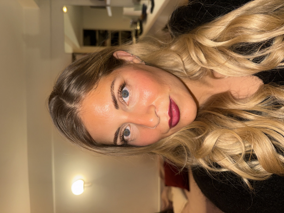A young woman with blonde, wavy hair is shown in a close-up shot. She has fair skin, light blue eyes, and dark red lipstick. She is wearing a black top. The background includes a neutral-colored wall and a glimpse of a room interior with a red pillow. The image appears to be taken indoors with warm lighting.