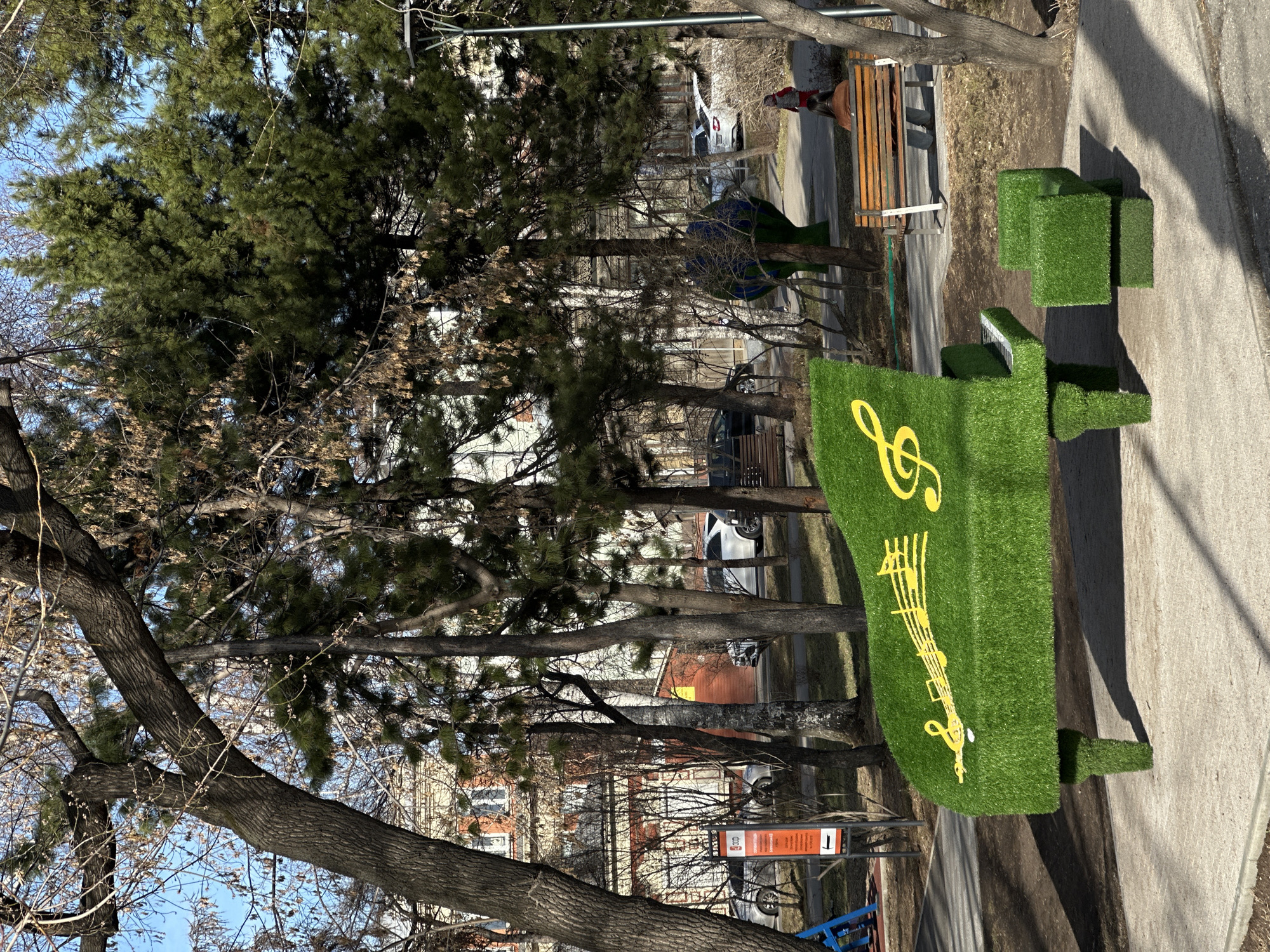 The image shows a city streetscape in Irkutsk, Russia. The central focus is a decorative, grass-covered sculpture resembling a grand piano, complete with a yellow treble clef and staff lines. Alongside the piano sculpture are three grass-covered cubic forms, likely part of the artistic arrangement. Behind the sculpture, there are several leafless trees interspersed with evergreen trees and a bench. The background includes parked cars and residential buildings. A street sign is visible. The street is paved and appears to be a pedestrian walkway. The lighting indicates it is daytime.