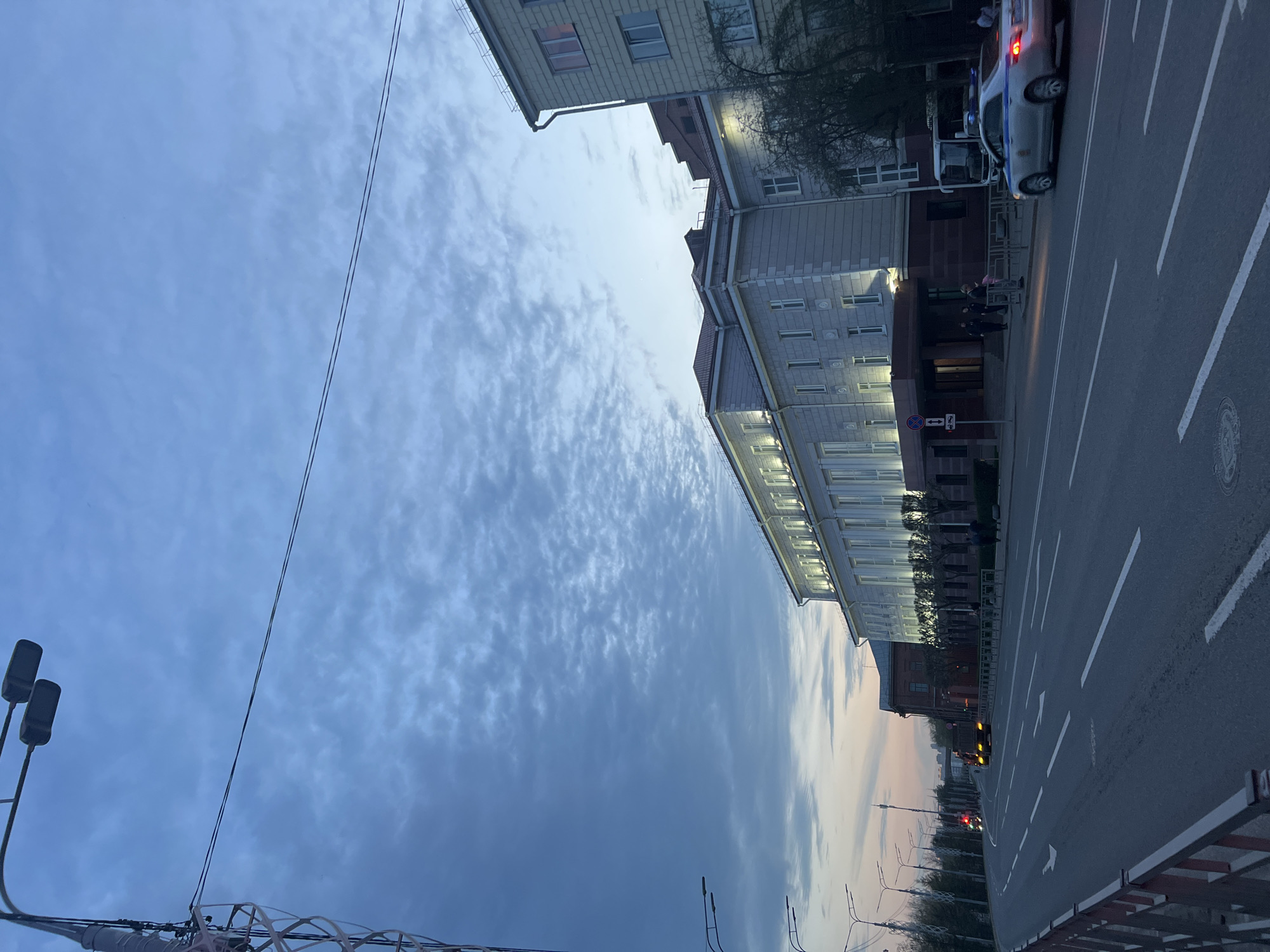 The image depicts a street scene at dusk in Krasnoyarsk, Russia. The sky is a mix of blue and grey with scattered clouds. A long building with white facade and evenly spaced windows runs along the right side of the street. A police car with its lights on is parked in front of the building, and a group of people are gathered nearby, appearing to be interacting. A road with white lane markings leads away into the distance, where more buildings can be seen. Streetlights are visible along the road. On the left, there are utility poles and power lines extending towards the sky. The overall environment suggests a city street with ongoing activity at the location of a building with a law enforcement presence.