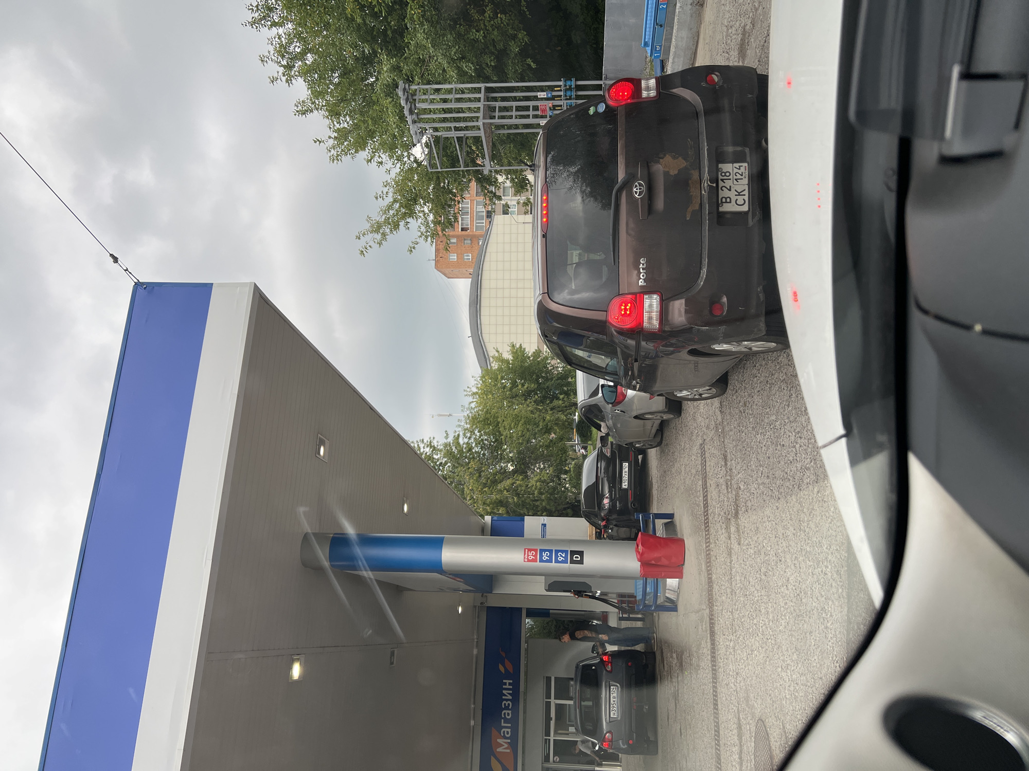 The image depicts a queue of cars at a gas station in Krasnoyarsk, Russia, under an overcast sky. In the foreground, the dashboard and windshield of a white car are visible. Further back, a brown Toyota Porte is parked, its brake lights illuminated. Behind it, other vehicles are lined up. A gas pump is present, with a person attending to it. The gas station features a blue and white canopy, with a sign displaying "Магазин" (Shop) in blue lettering. Trees and buildings are visible in the background, indicating an urban or suburban setting. The ground appears to be wet, suggesting recent rain.
