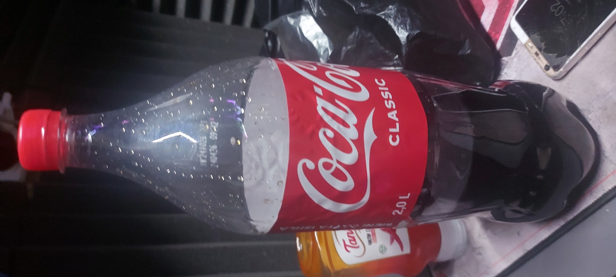 A partially-full 2.0L bottle of Coca-Cola Classic lies horizontally on a surface, with a red bottle of "Tanla" chili sauce visible underneath. The Coca-Cola bottle has a red cap and condensation droplets on its transparent section. A black plastic bag and a cracked-screened smartphone are visible near the Coca-Cola bottle. The scene appears to be indoors. The "Tanla" chili sauce indicates a possible location in Uzbekistan, given that "Tanla" is a brand produced there, likely in Tashkent.
