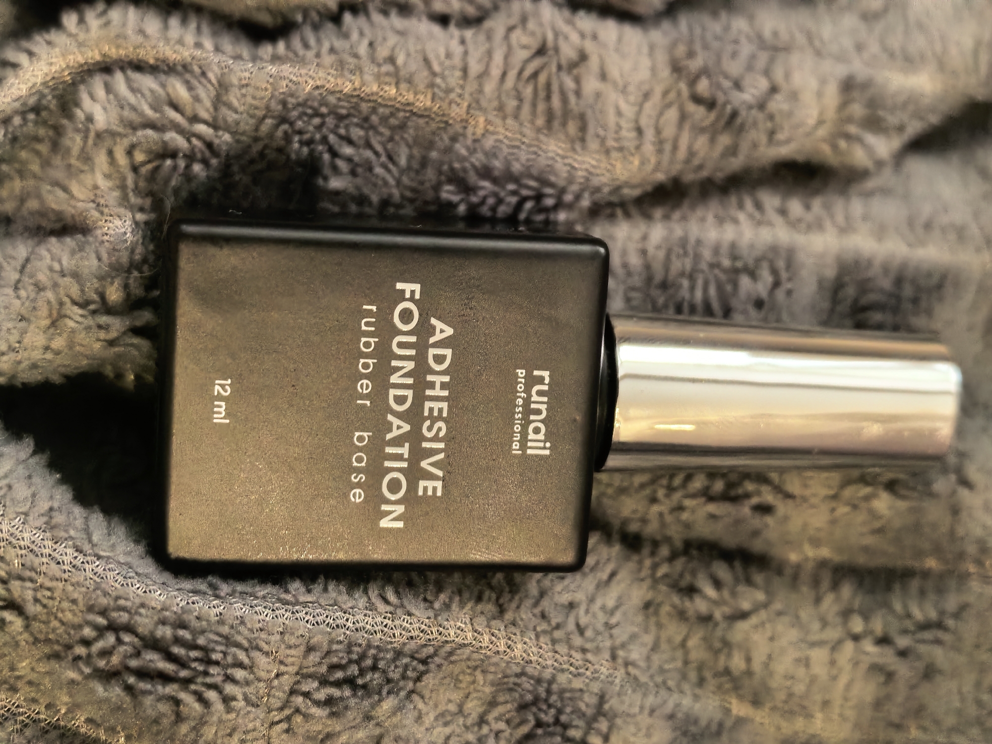 The image shows a bottle of "Runail Professional" adhesive foundation with "rubber base" written on the bottle. The bottle is rectangular, black, and lying on a grey, textured surface resembling a towel or blanket. The cap appears to be silver. "12 ml" is also printed on the bottle. The location is not specified in the image.