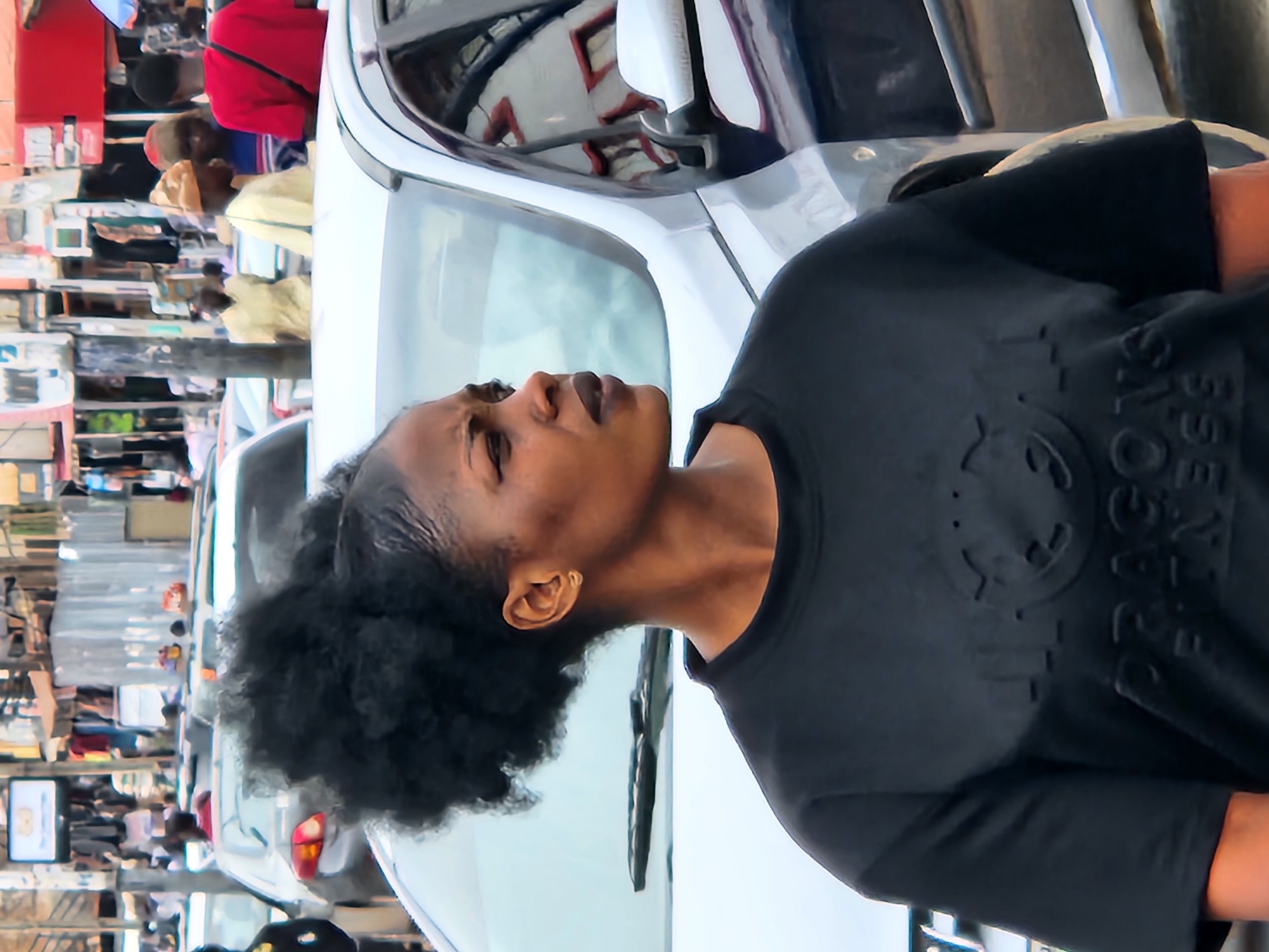 The image depicts a close-up of a dark-skinned woman in an outdoor, urban setting. The original image is rotated 90 degrees clockwise, showing the woman's head tilted towards the left, looking upwards. She has dark, curly hair pulled back, and is wearing a black t-shirt with embossed details.
On the right side of her chest, the text "DRAGONS BOYS" is clearly visible in an embossed, possibly embroidered, style. There is also a circular embossed pattern on the left side of her chest, which could be a logo or symbol. Her expression is neutral to contemplative, as she gazes upwards. A single gold earring is visible in her left ear.
Behind the woman, a white car is prominently featured, with its side and rearview mirror visible. The mirror reflects what appears to be part of a building or another vehicle with red and white elements. Further into the background, a bustling street scene unfolds. Several people are visible, including one person in a red top and another in a light yellow garment or head covering. These individuals appear to be walking or standing amidst a collection of buildings or market stalls. Hints of other vehicles and shopfronts with various items are also discernible, suggesting a busy commercial or residential street. The scene appears to take place during daylight hours, under bright, possibly sunny, conditions.