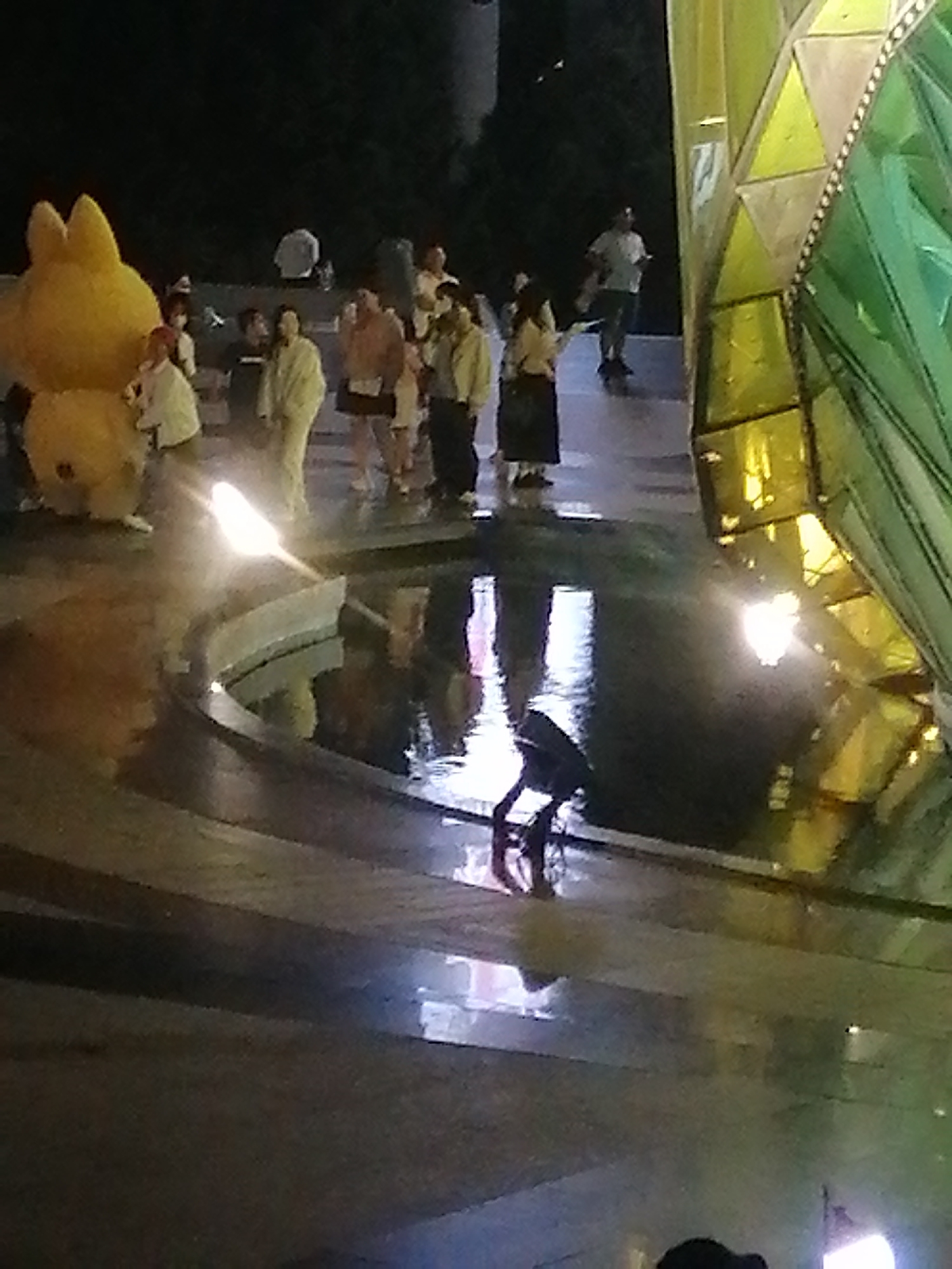 The image captures an outdoor evening scene, likely a public space with a water feature. In the foreground, the wet, reflective surface suggests it might have recently rained or the water feature is active. A bright light emanates from a curved structure in the water, illuminating the surrounding area and creating reflections. To the left, a large, yellow, cartoonish figure, possibly a mascot or inflatable, stands prominently. A group of people are gathered behind this figure, dressed in casual attire. Some are observing the scene, while others appear to be conversing. The background is dark, with hints of trees and structures, indicating an urban environment at night. In the mid-ground to the right, a large, geometric structure with yellow and green translucent panels is visible, adding a modern architectural element to the scene. Further back, a lone individual is walking. The overall impression is that of a lively public gathering space at dusk or night.