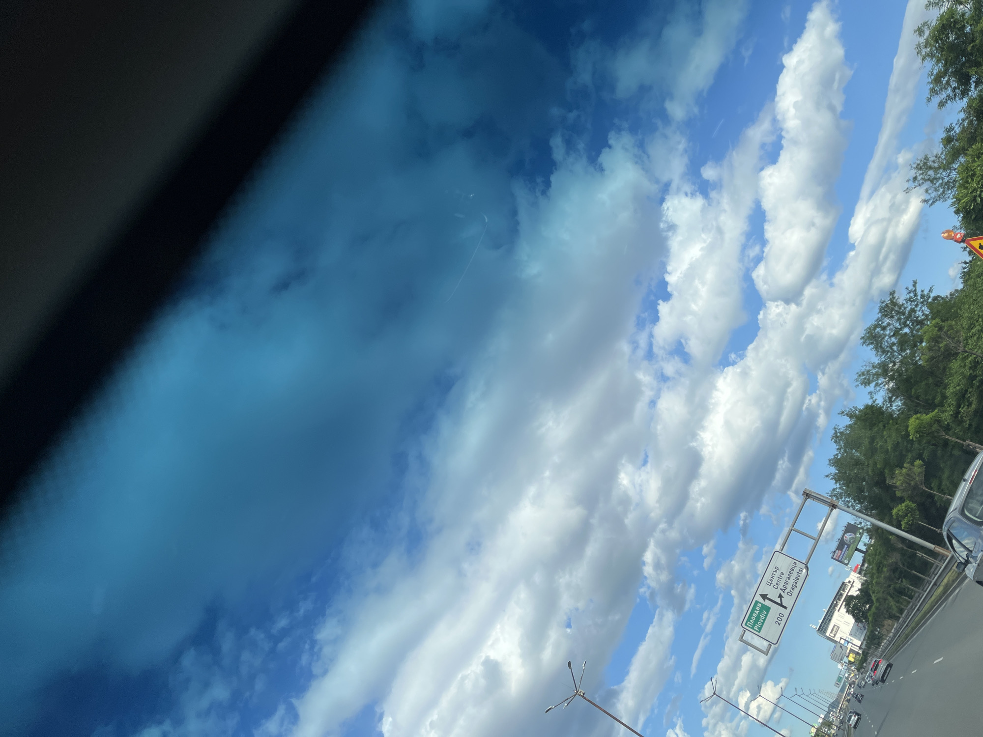 The image shows a view from inside a vehicle on a highway. The sky is filled with white clouds against a bright blue sky, with a tinted portion of the windshield obscuring the upper right corner. A road sign with directional information is visible on the right side of the highway. The sign is in Cyrillic and English, indicating directions for "Plovdiv Centre" and "Dragalevtsi," with distances noted. Several cars are driving on the highway, and trees line the side of the road. In the background, buildings and infrastructure suggest an urban or suburban environment.
