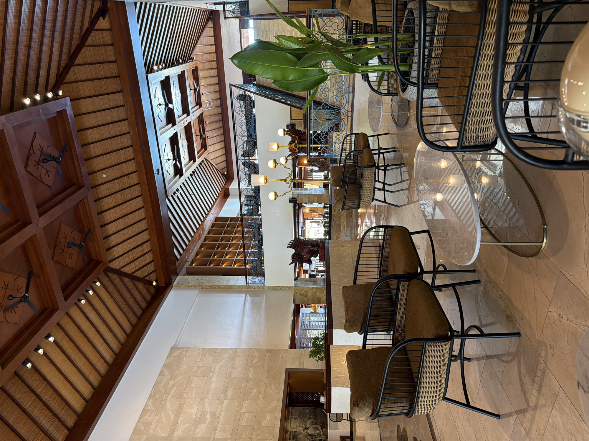 The image depicts an interior space, likely a hotel lobby or lounge, in Kuta, Indonesia. The architecture features a high, sloping wooden ceiling with decorative panels and recessed lighting. A second-floor mezzanine with ornate ironwork railings overlooks the lower level. The decor is a mix of modern and traditional Indonesian styles, including a large chandelier, wooden carvings, and a tall shelving unit filled with objects. Several armchairs with woven backs and dark metal frames surround small round tables. A large potted plant adds a touch of greenery. The floor is tiled in light beige. The overall scene suggests a luxurious and welcoming environment.