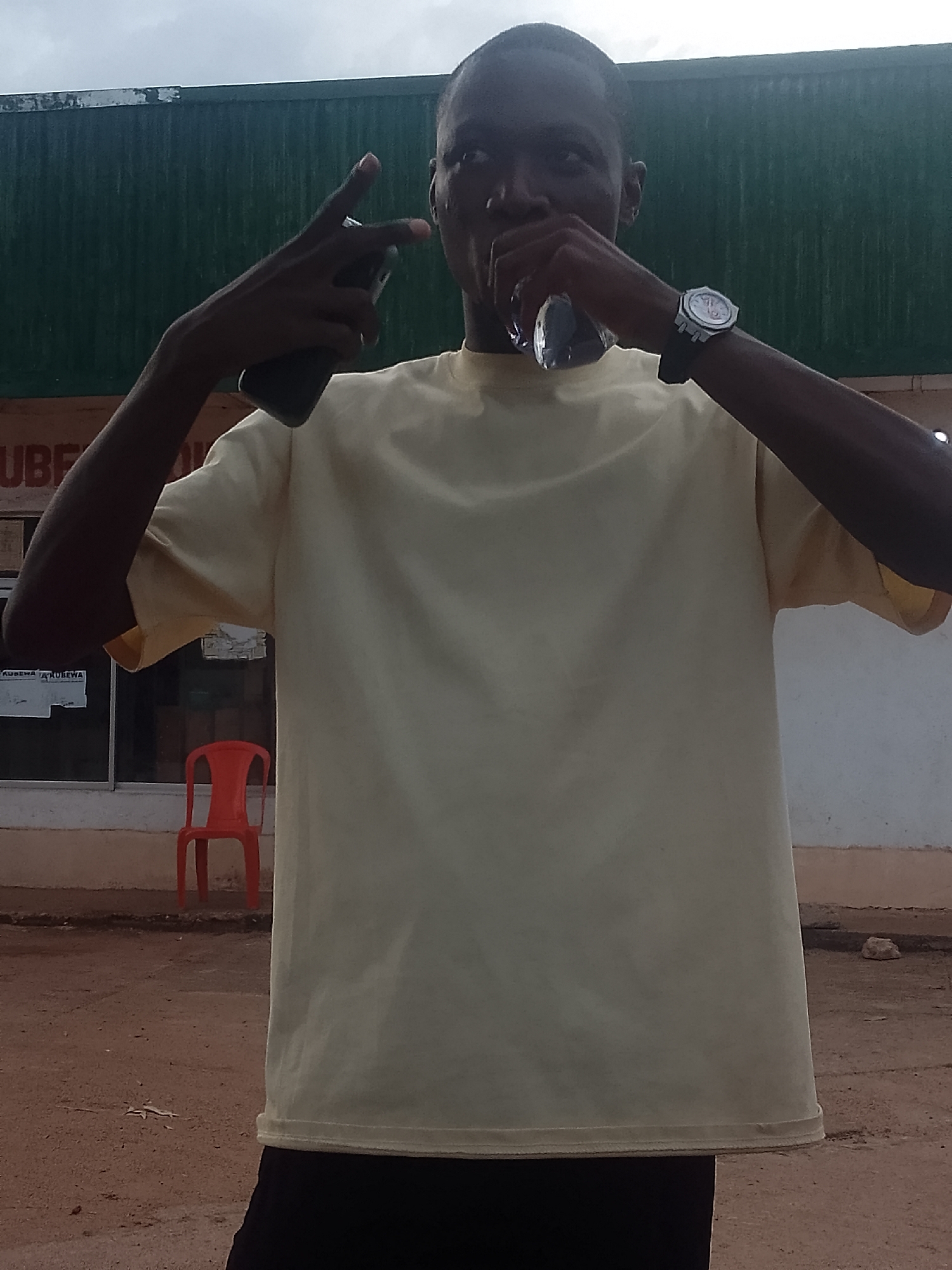 A young dark-skinned man stands on a dirt surface in front of a building, making gestures with his hands. He is wearing a light-yellow t-shirt, black pants, and a wristwatch on his left wrist. In his right hand, he holds a dark-colored cell phone, gesturing with his fingers. An orange chair is visible behind him, near a window. The building has a green corrugated metal awning. The location is Unknown, Unknown.