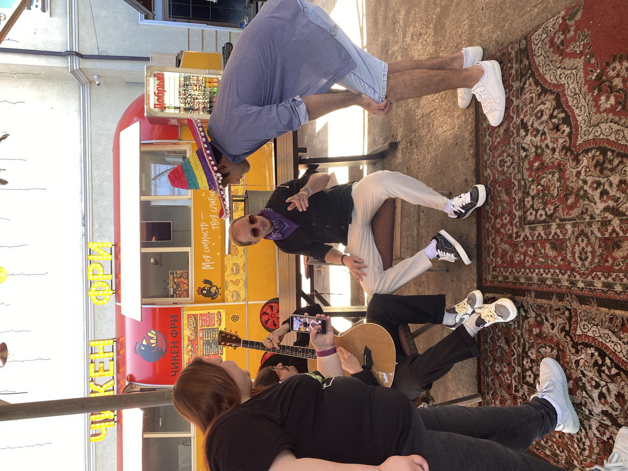 This media file captures a vibrant, casual outdoor gathering, likely in a cafe or food truck setting in Sochi, Russia, given the Cyrillic text and location context. The scene is brightly lit, indicating daytime.
Several people are present, engaged in a social interaction. A man in a blue polka-dotted shirt, denim shorts, and a colorful sombrero leans forward, seemingly interacting with a seated man. This seated man wears a black shirt, white pants, a purple bandana, and sunglasses, and is pointing playfully. Another individual is seated, holding an acoustic guitar, suggesting a performance or jam session. Next to them, a person holds a smartphone, apparently recording the guitar player or the general scene. In the foreground, a woman with reddish-brown hair is visible from behind. Various feet in different sneakers are seen on the concrete ground and an ornate red and beige patterned rug.
A prominent red food truck with yellow accents serves as a backdrop. Its signage includes glowing yellow letters spelling "ЧИКЕН ФРИ" (Chicken Fry). Other visible Cyrillic text reads "ЗАКАЗ" (Order), "КОМБО ЧИКЕН ФРИ" (Combo Chicken Fry), "Моя сочность" (My juiciness), "твоя слабость" (Your weakness), and "Добрый" (Good/Kind), along with a phone number. The truck also features a cartoon chicken logo. The overall atmosphere appears relaxed and friendly, portraying a group enjoying music and each other's company.