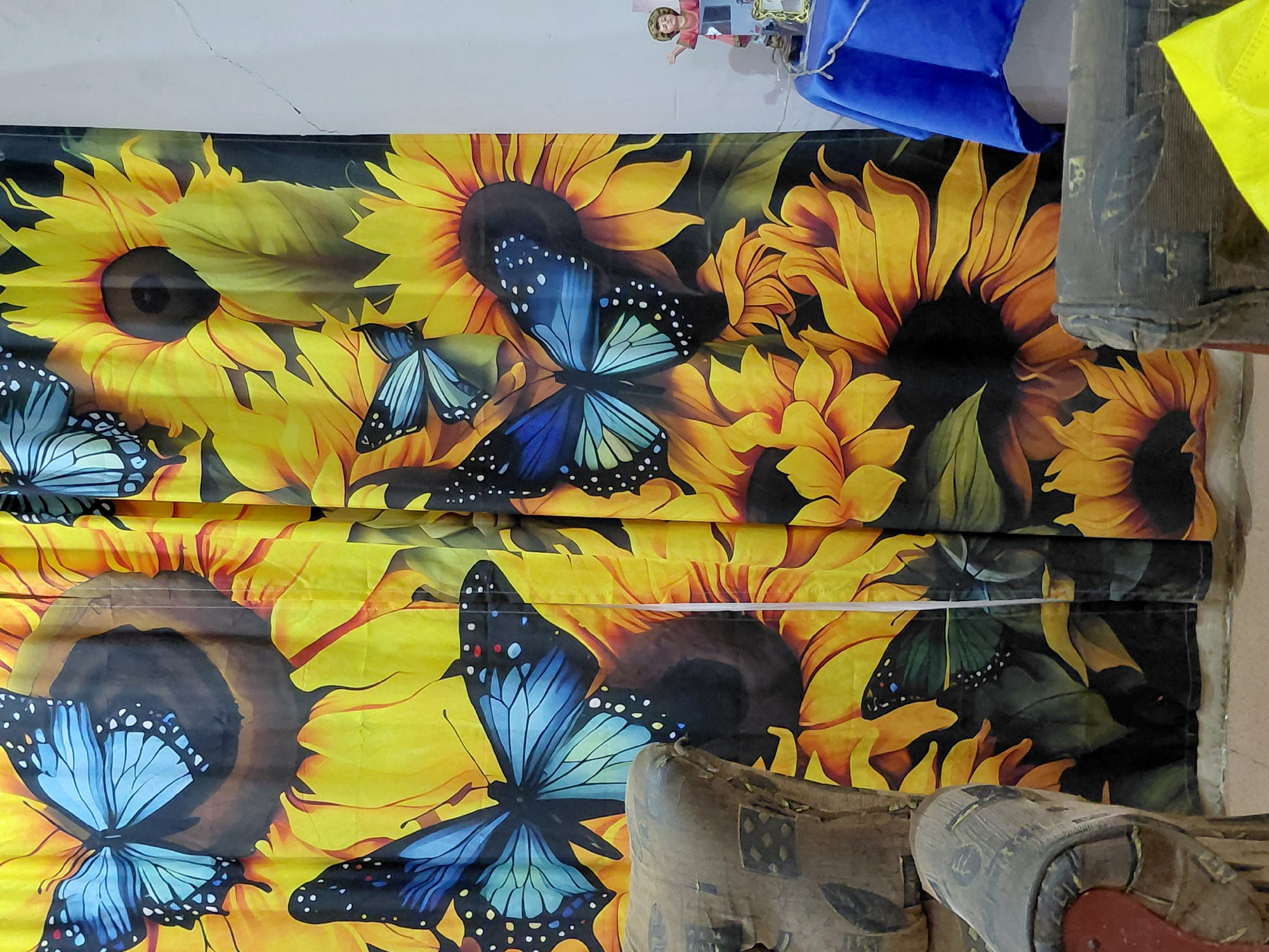 The image depicts a close-up of a yellow curtain with a pattern of sunflowers and blue butterflies, hanging vertically. The curtain appears to be in a room with two upholstered chairs partially visible on the left and right sides of the frame. The chair on the left is brown and shows signs of wear, with some fabric torn. The chair on the right is also partially visible and appears to be made of a darker material. To the far right, a small statue of a child in red clothing with arms outstretched is visible on a table covered with a blue cloth. A white cord or string is draped across the table. The background wall is white and shows some cracks. The scene is likely an indoor setting, possibly a living room or a bedroom. The specific location in Santa Ana, Venezuela is not determinable from the visual information provided.