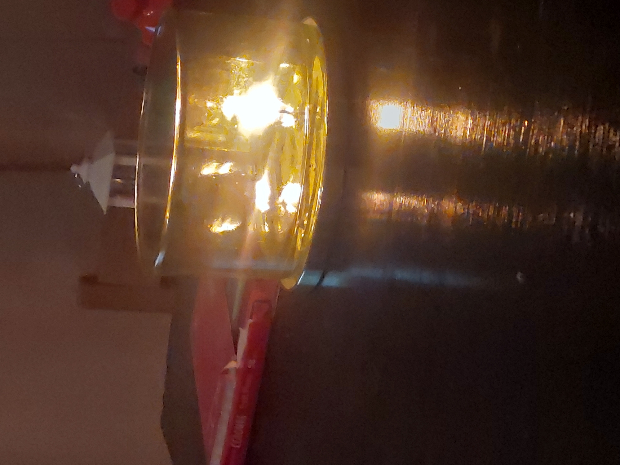 The image shows a lighthouse-style table lamp with a bright, golden glass shade. The lamp sits atop a red book or object on a dark, possibly wooden, surface. The wall behind the lamp appears light beige. The reflective light on the dark surface suggests the lamp is indoors. There is no direct indication of the location being Tsarevo, Bulgaria.