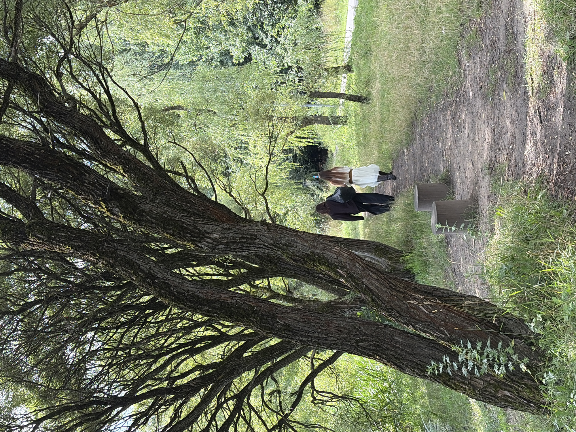 Two individuals are walking away from the camera on a dirt path in a park. The person on the left is wearing dark clothing, including pants and a long-sleeved shirt, and carrying a dark bag. The person on the right is wearing a white dress, dark boots, and has long brown hair. They are walking under a large tree with extensive branches. The path is bordered by grass and other trees, including weeping willows. The scene appears to be in a natural setting, possibly a park or wooded area. The location is identified as the Park of Levoberezhny in Khimki, Russia.