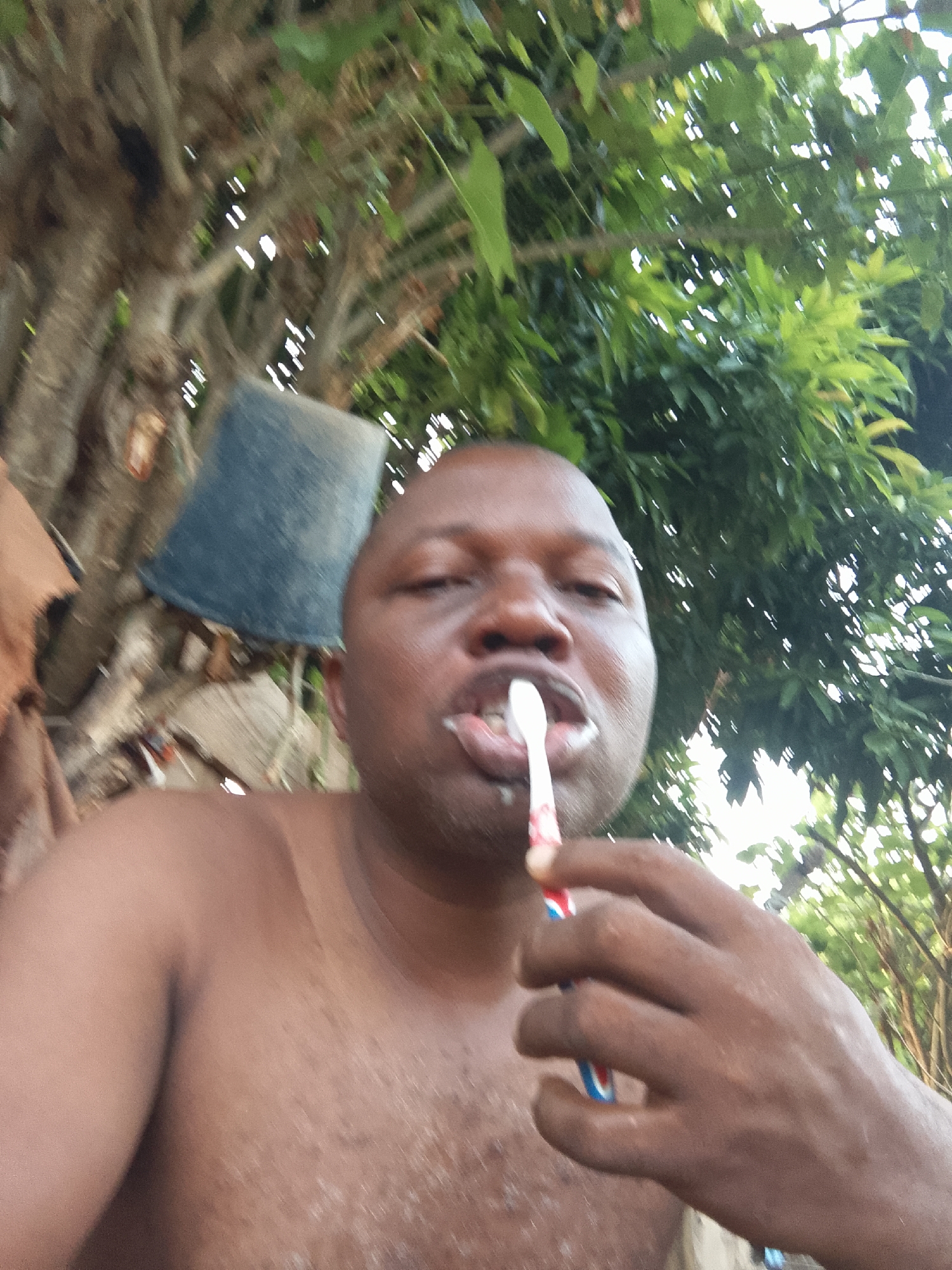 A dark-skinned, bald man is brushing his teeth outdoors. He is shirtless and appears to be holding the toothbrush in his right hand. The background includes a tree with green leaves and what appears to be a blue bucket or container hanging from it. The location and city are unknown.