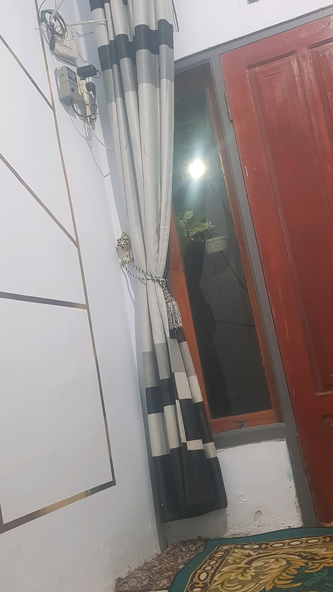 The image shows an indoor scene with a white wall, a curtain, a window, and a red door. On the white wall, there is an electrical outlet with wires. The curtain is grey and black striped and tied back with a decorative tassel. The window is dark and reflects a light source. The red door is on the right side of the image. A patterned rug is visible on the floor. The scene appears to be located in Bakalan, Indonesia, likely in a residential setting, judging by the domestic objects present. There are no people visible in the image.