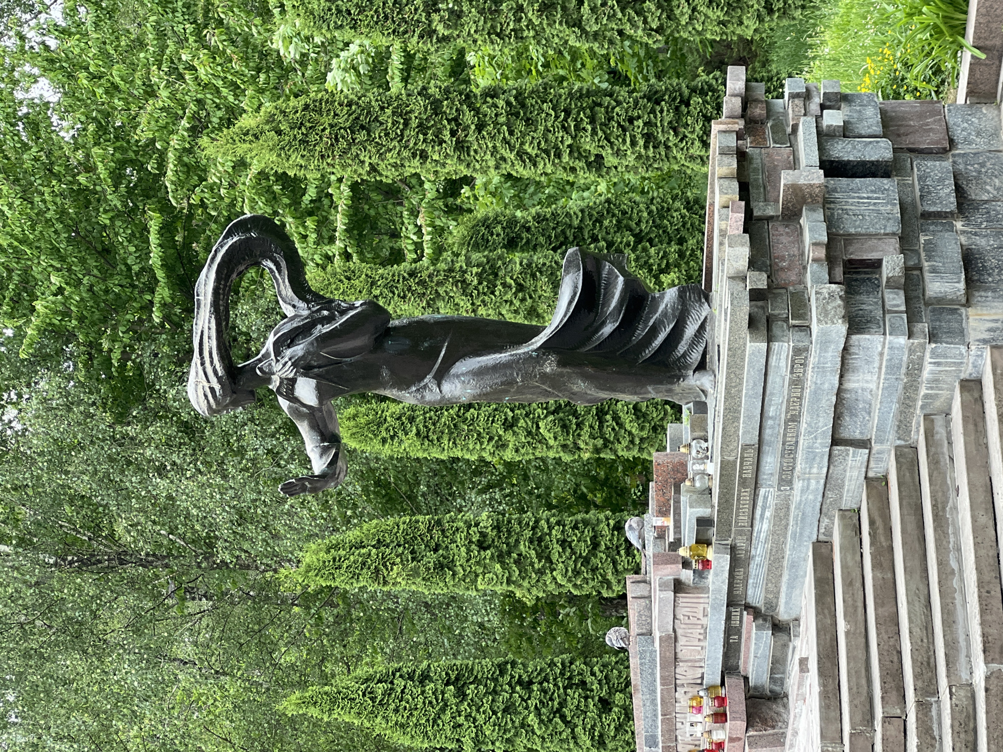 The image shows a sculpture titled "The Unbending," dedicated to the Victims of Political Repression in Ternopil, Ukraine. The statue of a female figure with flowing hair is set against a backdrop of green trees and foliage. To the side of the statue is a multi-tiered memorial made up of stone slabs or blocks, likely containing names or inscriptions. The slabs are of various colors and textures. There are also several candles and small decorative objects placed on the memorial.