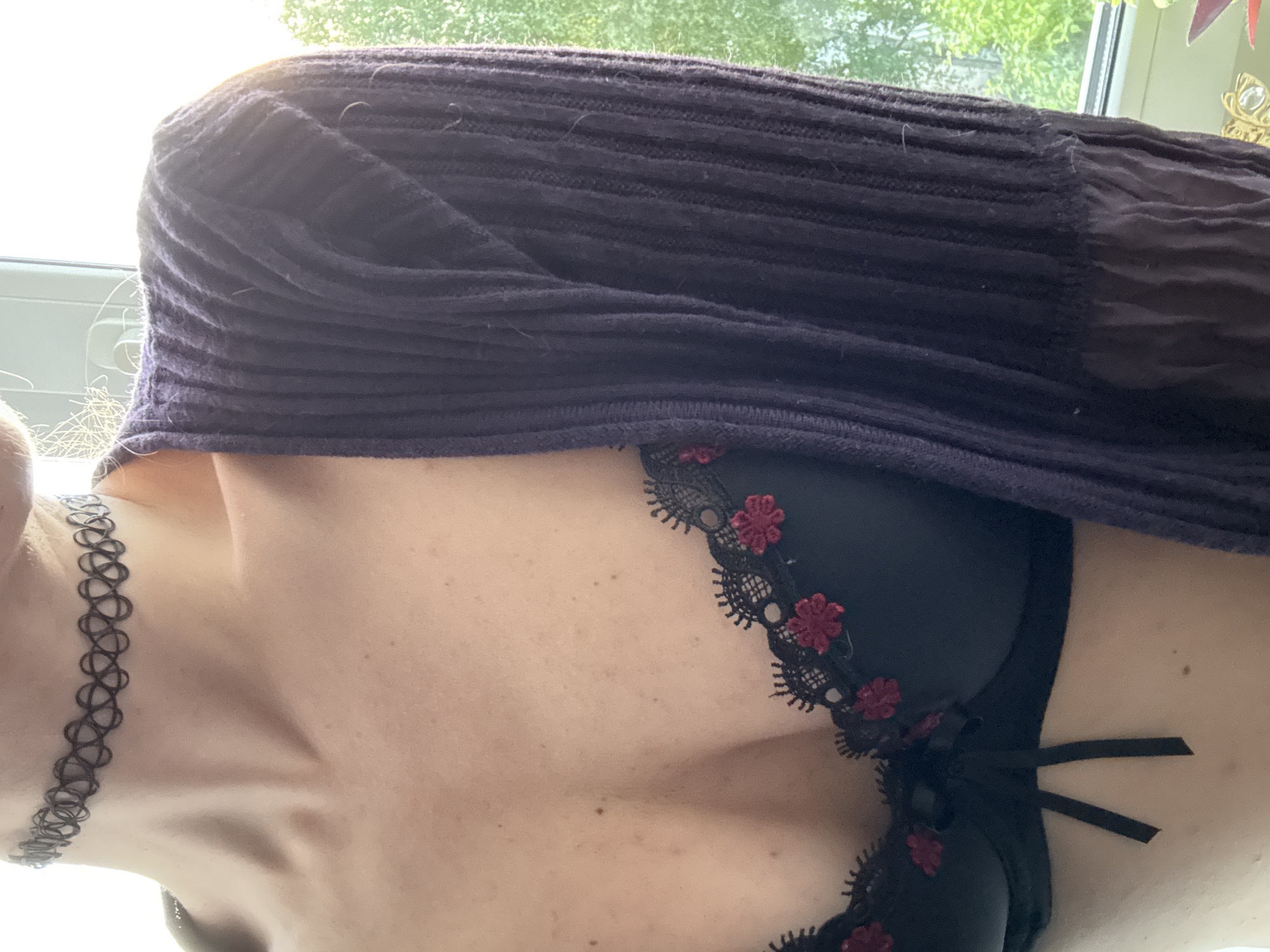 The image shows a close-up view of a person's upper body, specifically their chest and neck. They are wearing a black bra with red floral lace detailing and a dark purple knitted cardigan. A black tattoo choker necklace is visible around the neck. The background appears to be a window with some greenery outside, suggesting an indoor setting, possibly in Chaikovsky, Russia, though this cannot be confirmed from the image alone. The lighting suggests it might be daytime.
