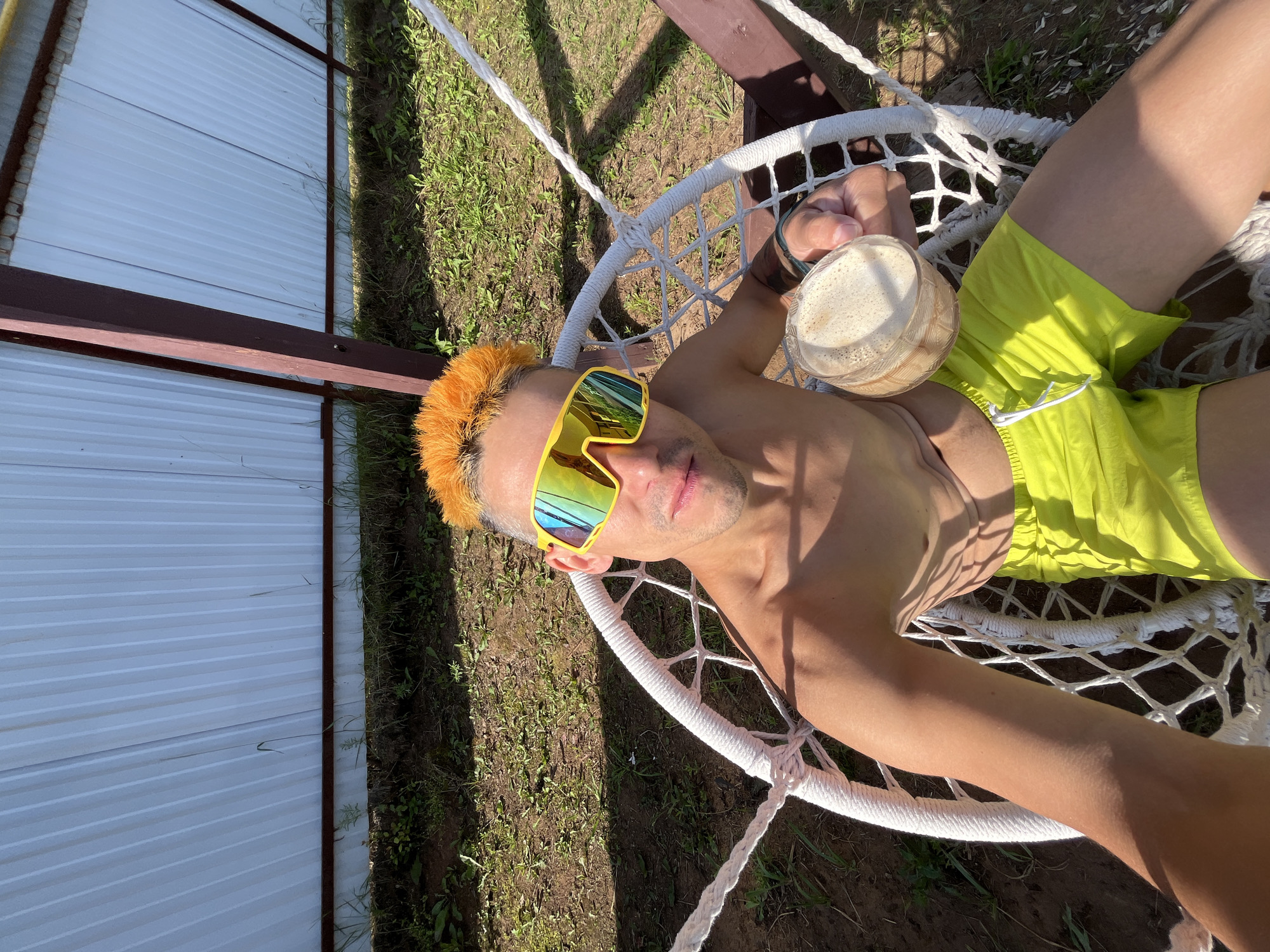 A person with bright orange, spiky hair and yellow, reflective sunglasses is lying on a white, macrame hammock. They are wearing bright green shorts. The person is holding a clear glass with a frothy, light-colored drink in their left hand. The hammock is suspended outdoors, with grass and dirt visible beneath it. Sunlight casts shadows from above, suggesting it's daytime. To the left of the hammock is a white corrugated metal wall, possibly part of a building or fence, with a dark brown wooden beam running horizontally across it. The overall scene appears to be a relaxed, summery setting. The individual's pose and the drink suggest they are enjoying leisure time.