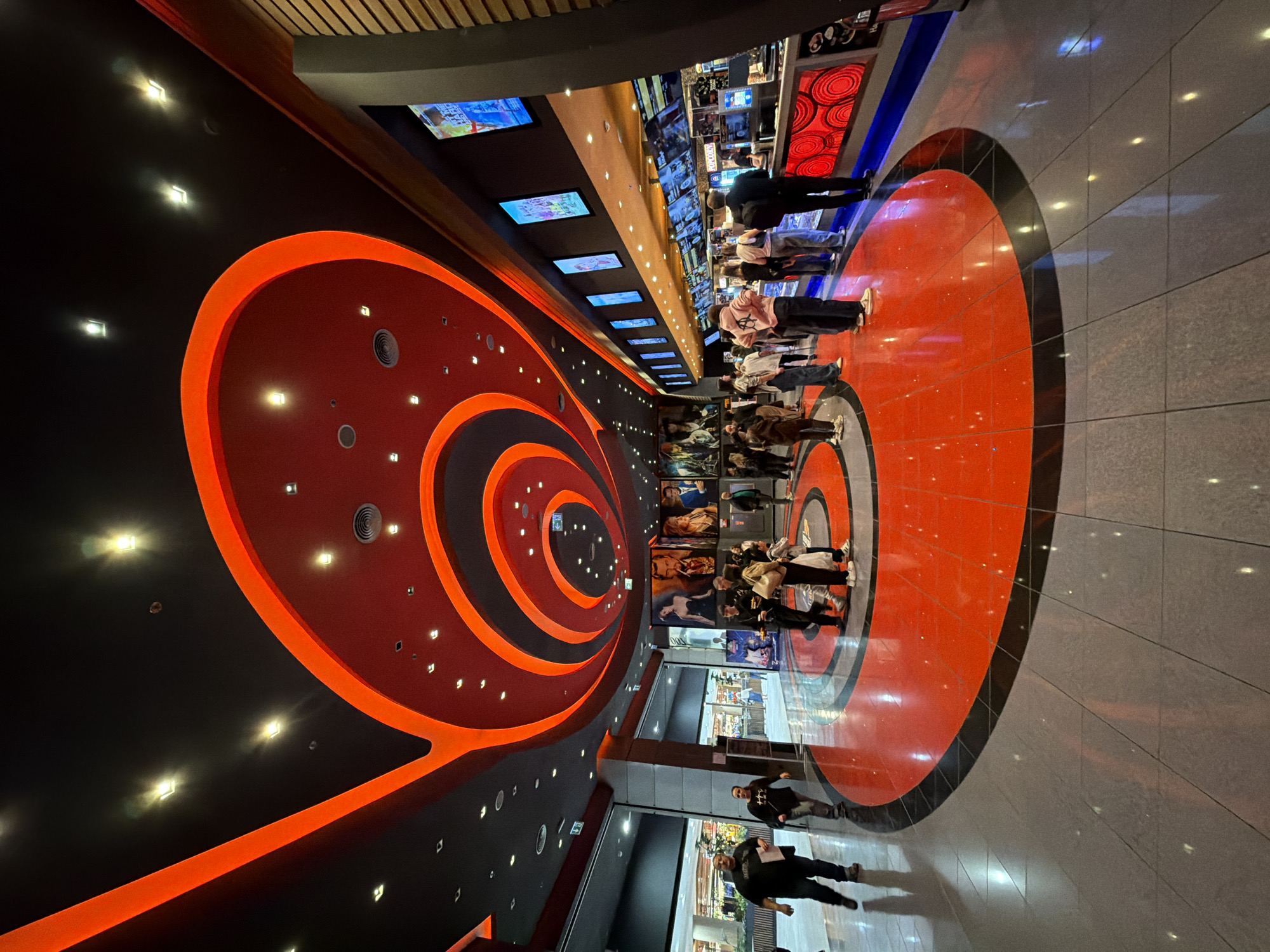 The image captures the interior of the Arena Cinema complex in Sofia, Bulgaria. The scene showcases a wide corridor with a unique circular, spiral design pattern on the ceiling, featuring alternating bands of red and black with integrated lighting. People are present in the corridor, some appearing to be waiting in line, while others are walking through the space. Movie posters and monitors displaying film trailers line one side of the corridor. The floor design mirrors the spiral motif of the ceiling, with red and black circular segments extending to the adjacent gray tiled flooring.