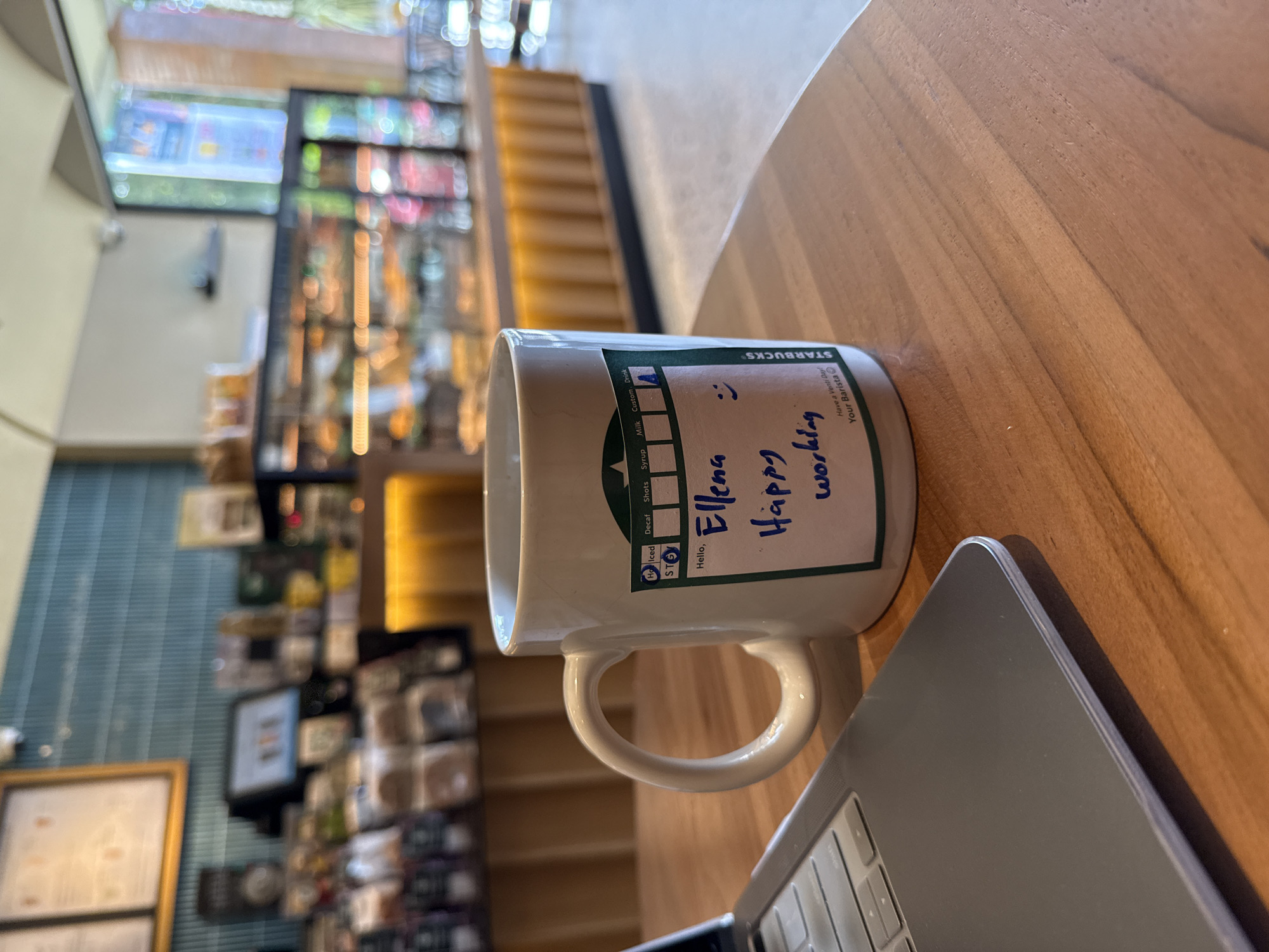 A white Starbucks mug with a handwritten message, "Hello, Ellena, Happy working," sits on a wooden table next to a laptop. The mug has the Starbucks logo and a white sticker with handwritten text and a smiley face. The background shows the interior of a Starbucks coffee shop, with shelves of products and a counter area, suggesting it is located in a public space like the one in Kuta Selatan, Indonesia.