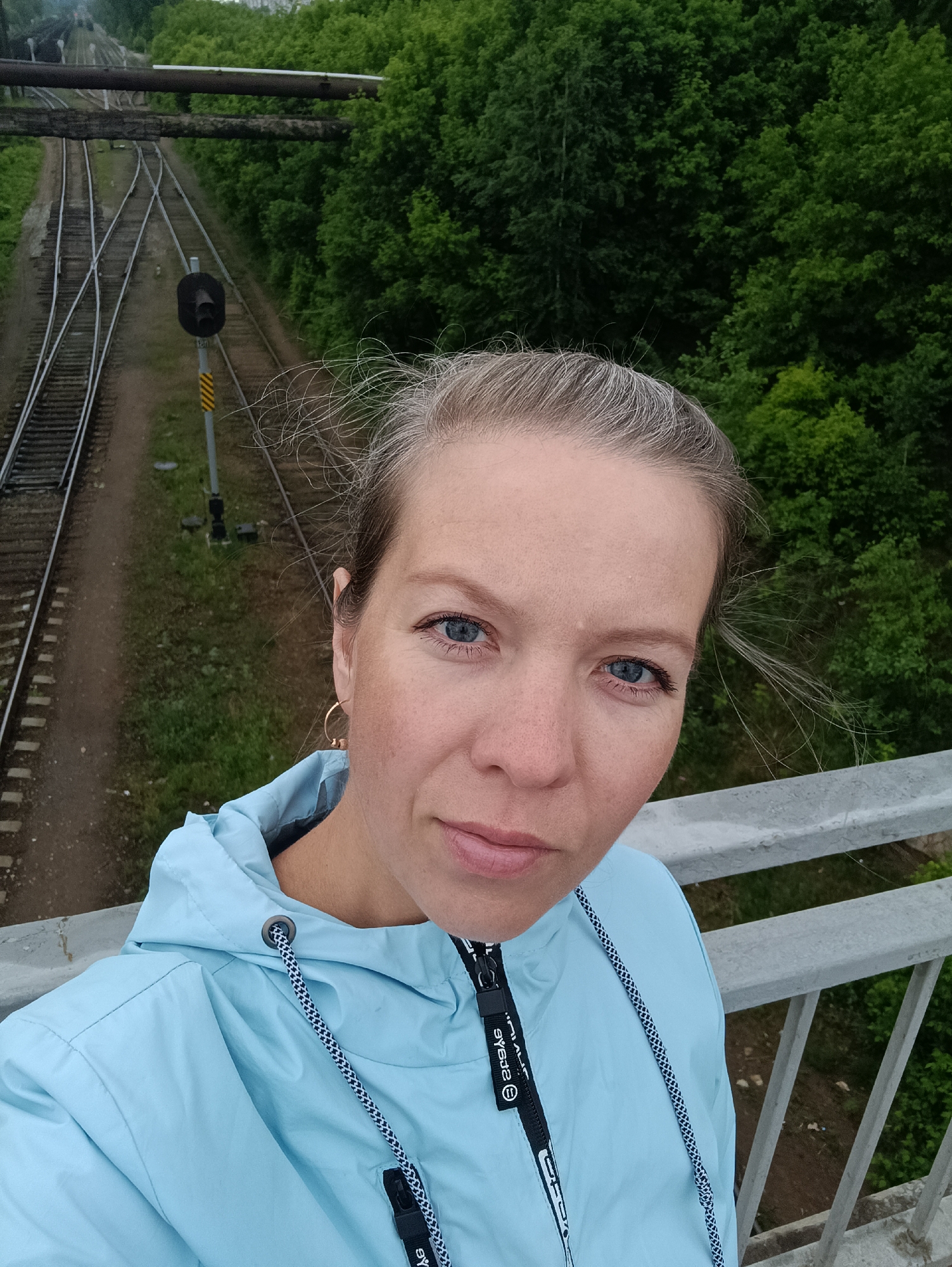 The image is a selfie of a fair-skinned woman with blonde hair and blue eyes. She is wearing a light blue jacket and gold hoop earrings. The woman is positioned on a bridge or elevated structure, as evidenced by the grey metal railing in the foreground.
In the background, there are railroad tracks with switching points, suggesting a railyard or station. A railway signal post is visible next to the tracks. Above the tracks, there appear to be pipes or conduits spanning across the scene. The environment includes lush green trees and foliage, suggesting the image was taken during a season with abundant vegetation.
The location is Chaikovsky, Russia.