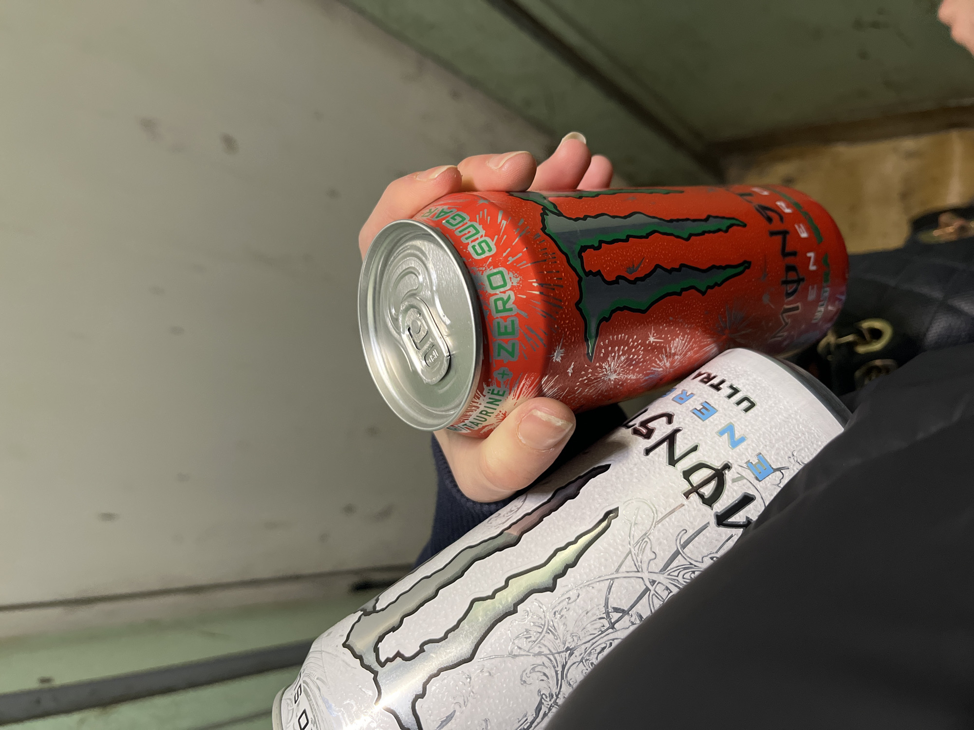 A close-up image shows a person's hand holding two Monster Energy drink cans in what appears to be an indoor or covered setting, possibly in Sofia, Bulgaria. The prominent can is a red Monster Zero Sugar, held upright with the silver pull-tab top facing the viewer. Green text reading "ZERO SUGAR" and "TAURINE" is visible on its side, along with the green Monster logo and a design featuring white sparks or fireworks against the red background. Condensation covers the surface of the can, indicating it is cold.
Partially visible beneath the red can and held by the same person, or resting nearby, is a white Monster Ultra Energy can. Its design includes the black Monster logo, intricate grey patterns, and the text "MONSTER ULTRA ENERGY" and "ULTRA" in black and light blue lettering. This can also exhibits condensation.
The person holding the cans is mostly out of frame, with only their hand and a small portion of a dark-colored garment (likely a jacket or sleeve) visible. To the right, a section of a black quilted bag with a gold-colored emblem or chain is also present. The background consists of plain, light grey or beige walls with some darker marks and indistinct features, suggesting a simple, functional environment such as a transport vehicle or public waiting area.