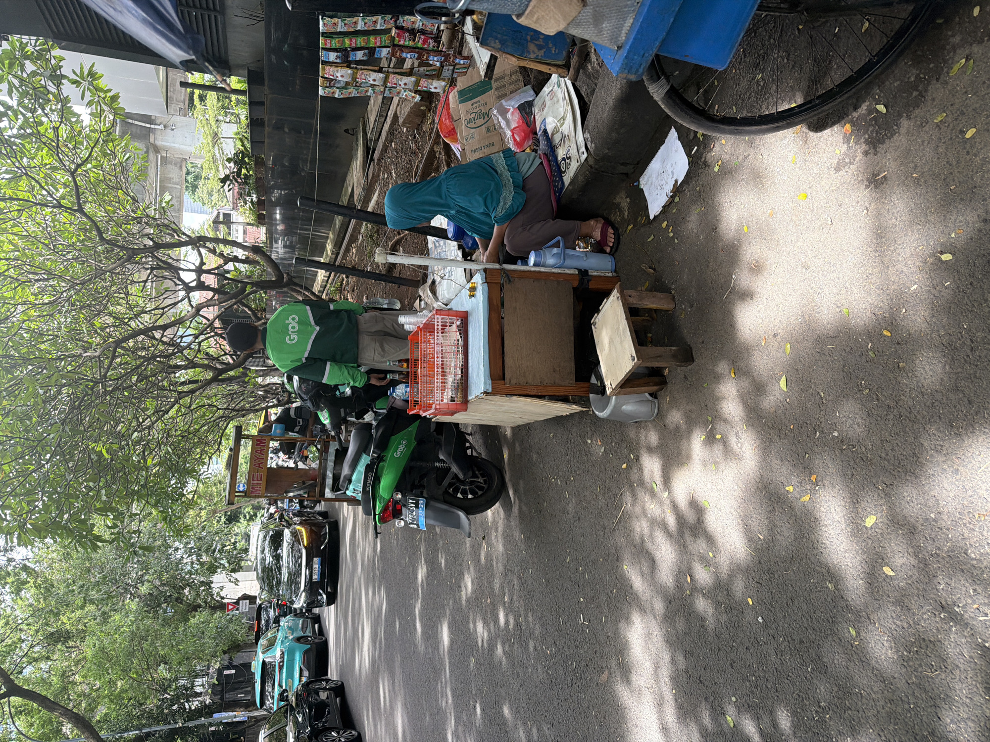 The image shows an outdoor scene in what appears to be a street in South Jakarta, Indonesia. The image is rotated 90 degrees. On the right, a woman wearing a hijab kneels next to a food cart. The cart appears to be mobile, possibly attached to a bicycle. The cart is cluttered with various items, including drinks, food packaging, and cooking utensils. A man is standing by the cart, wearing a uniform that says "Grab" which indicates he is a delivery driver. Several green Grab-branded motorcycles are parked along the street. The street is lined with cars and a few motorcycles parked on the side. Shadows of trees cast patterns on the road surface, suggesting a sunny day. The text on the food cart reads "Mie Ayam," which means chicken noodle. One can also see the Indonesian text "Buka Disini" and the brand name "Marjan." License plates of the vehicles are partially visible, further pointing to the location in Indonesia.