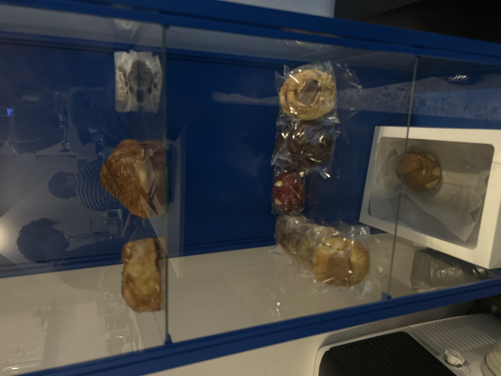 A display case with a blue frame contains assorted baked goods, including pastries and cookies, some individually wrapped in clear plastic. One unwrapped pastry is visible on the bottom shelf. A white cardboard container holds a single bun. Reflections visible in the glass of the case suggest a human figure wearing a striped shirt and another person with dark hair in the background. The scene appears to be indoors, with a white surface beneath the case and other objects partially visible at the bottom. The exact location, San Bernardino Tlaxcalancingo, Mexico, is not discernible from the image.