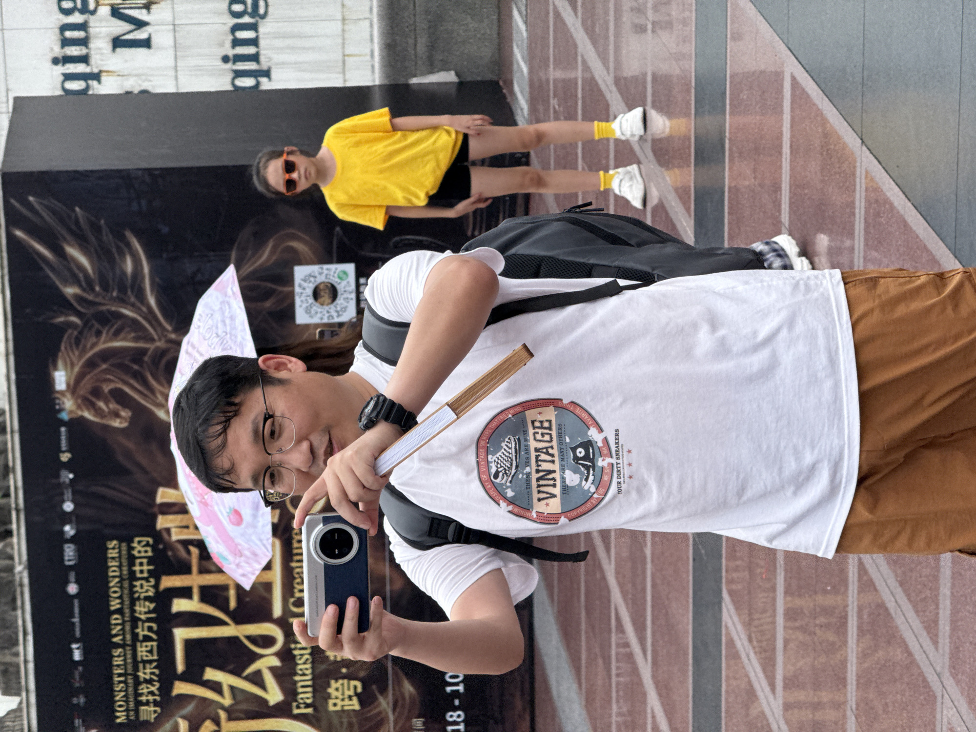 A man wearing glasses and a white t-shirt with "VINTAGE" printed on it holds a blue and silver camera to take a photo. He is holding a folded fan in his right hand. A young girl in a yellow t-shirt and black shorts stands in the background, wearing sunglasses. The scene takes place in a public square with patterned flooring, likely in Yuzhong District, China, with a large banner in the background featuring text in English and Chinese.
