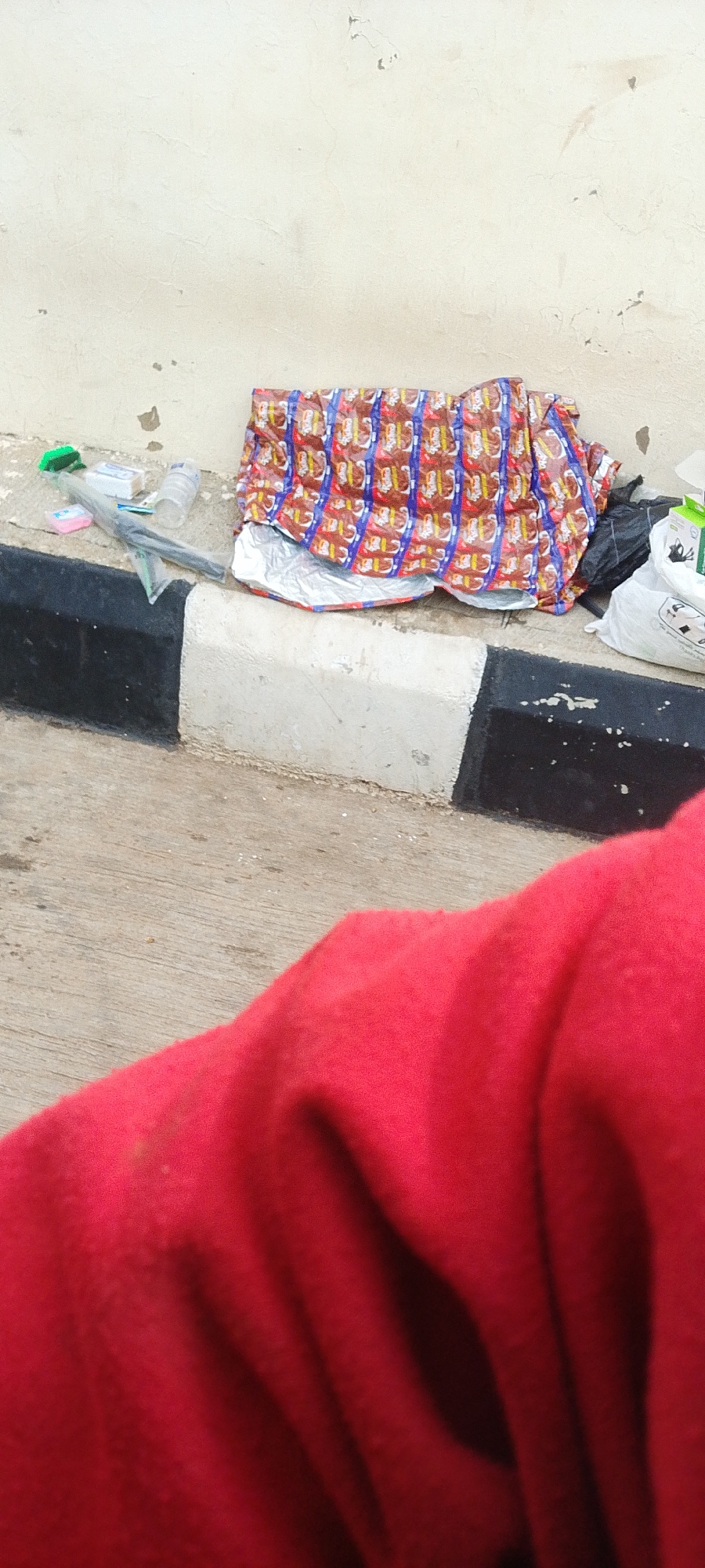 This image depicts a scene on a sidewalk in the city of Jos, Nigeria. In the foreground, a large portion of a red cloth, possibly a garment or blanket, is visible, obscuring much of the lower part of the frame. In the middle ground, various objects are scattered on the ground and on the curb. A striped curb, black and white, separates the sidewalk from the street. To the left of the curb, there are some small items including what appears to be a green brush, a small pink object, a bottle of water, and some plastic packaging. Dominating the center and right side of the middle ground is a large, colorful package or blanket with a repeating pattern of what looks like food items in red, blue, and yellow wrappers. Behind this, on the right side, are black and white plastic bags, one of which is a white shopping bag with some printed text. The background is a textured, light-colored wall, likely part of a building. The overall impression is one of discarded items on a public street.