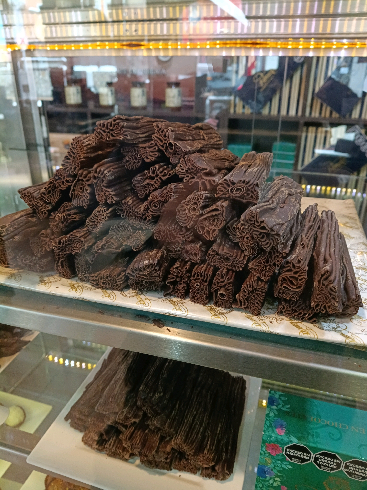 The image shows a display case filled with chocolate confections. In the foreground, a pile of dark chocolate pieces, textured and layered, are arranged on a patterned paper. These pieces resemble bark or twisted ribbons. Below this, in the lower portion of the frame, another pile of similar chocolate pieces is visible on a white rectangular plate. The display case features a metallic shelf and a glass front, reflecting the interior lighting and background elements. The background shows shelves with various items, some appearing to be jars and possibly other food products, suggesting a commercial establishment. A green box with floral designs and warning labels indicating excess sugar, total fat, and saturated fat is partially visible in the lower right corner. The lighting is bright, highlighting the chocolate products. The image does not provide any information or visual cues to associate it with Ushuaia, Argentina.