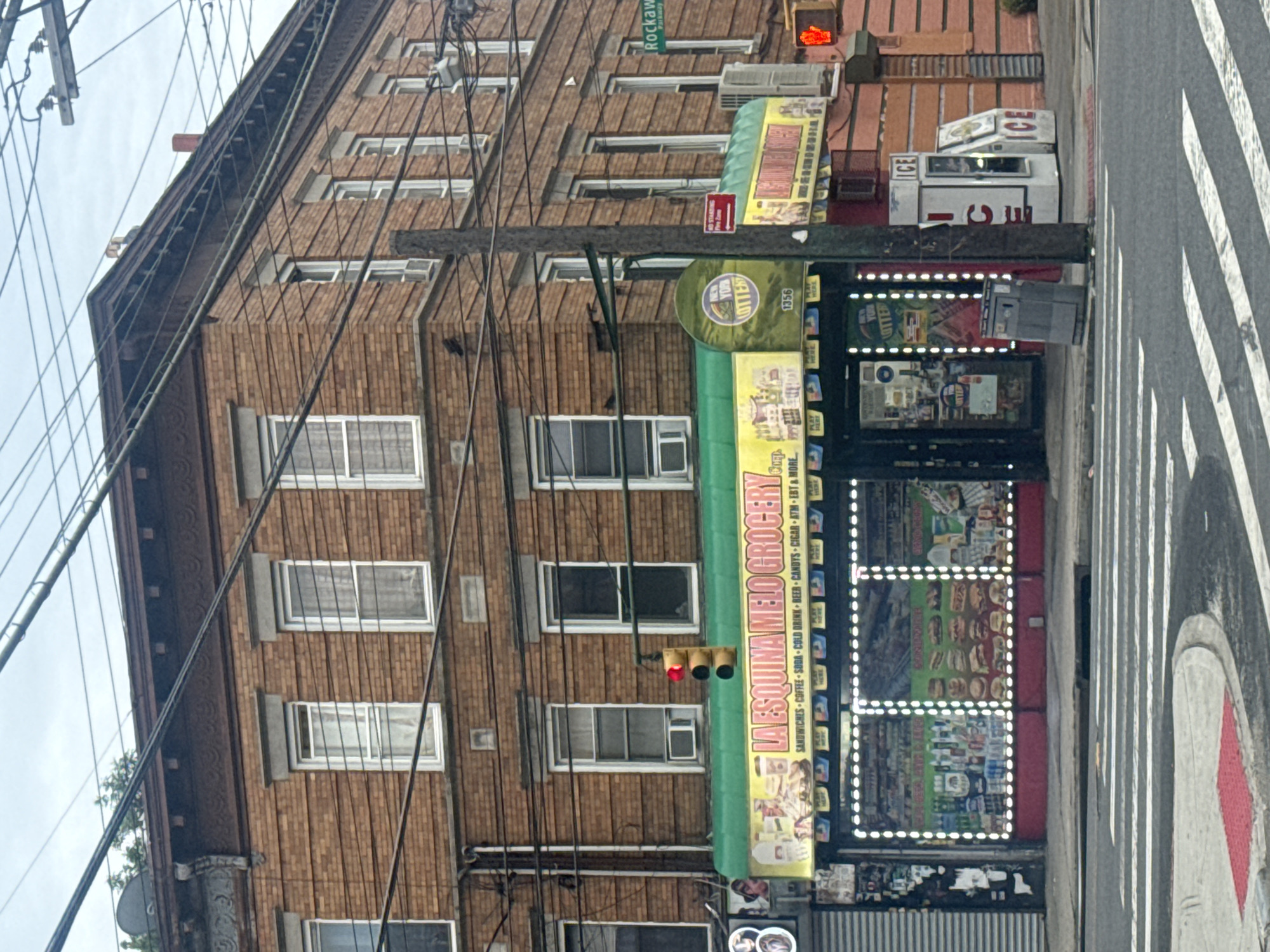 This is an eye-level, medium shot of a corner grocery store called "LA ESQUINA MELO GROCERY" in Queens, New York City, USA. The building is made of brick and has multiple windows on the upper floors. The storefront is decorated with bright lights and advertising for various products and services like sandwiches, coffee, soda, beer, candy, cigars, ATM, and EBT. There is a New York Lottery sign and the number 1356 is visible. To the right of the store, there are two ice vending machines. A traffic light shows a red light, indicating a stop. A pedestrian crossing is visible in the foreground. The sky is overcast.