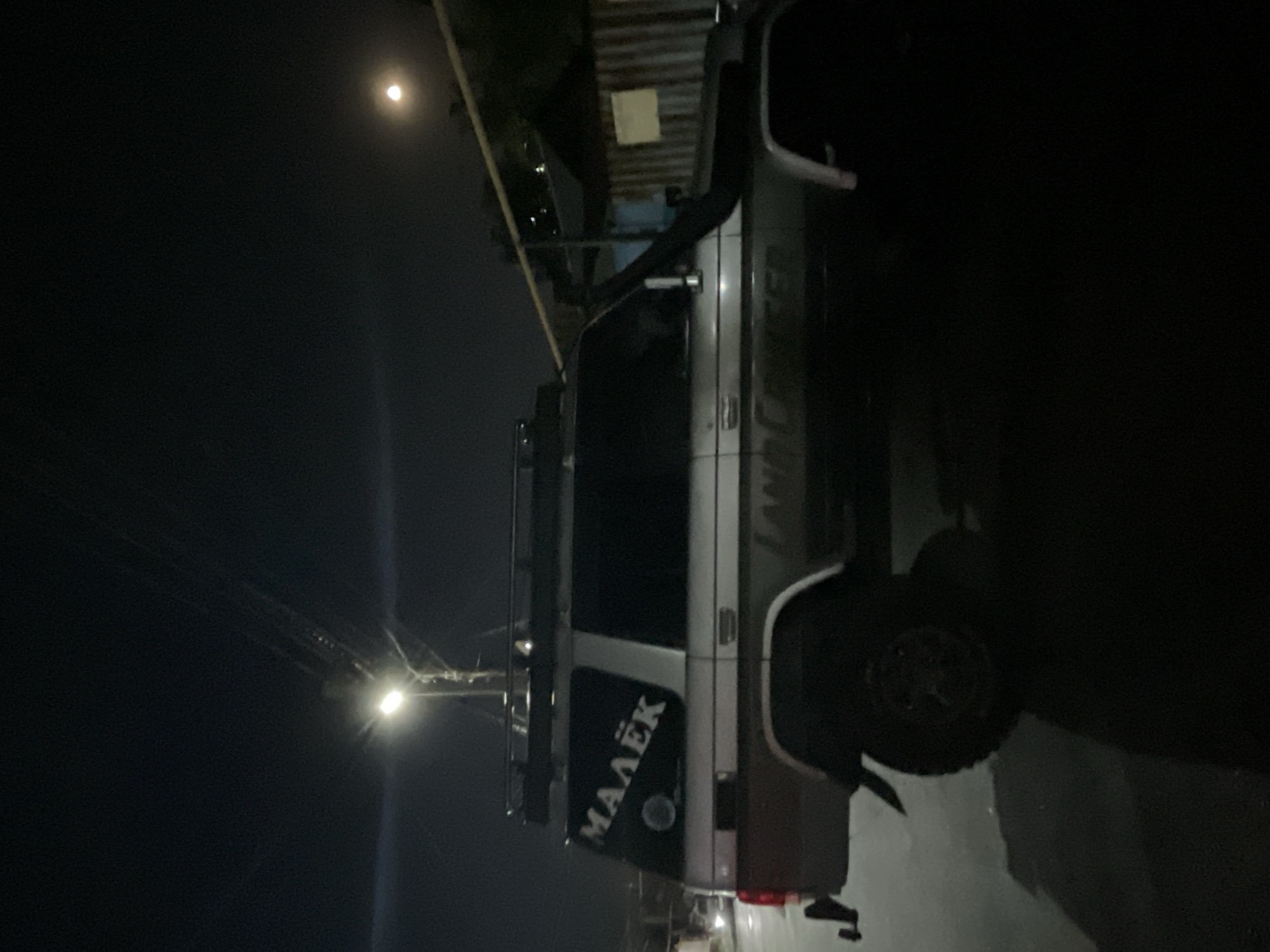 A side view of a silver Toyota Land Cruiser parked on a paved road at night. The vehicle has a roof rack and a snorkel on the driver's side. A sticker on the rear passenger window reads "МАЛЁК" in white Cyrillic letters. The street is illuminated by a single street lamp. In the background, there are buildings and a power line. The moon is visible in the upper right corner of the image. The scene appears to be in the city of Almaty, Kazakhstan, given the context provided.