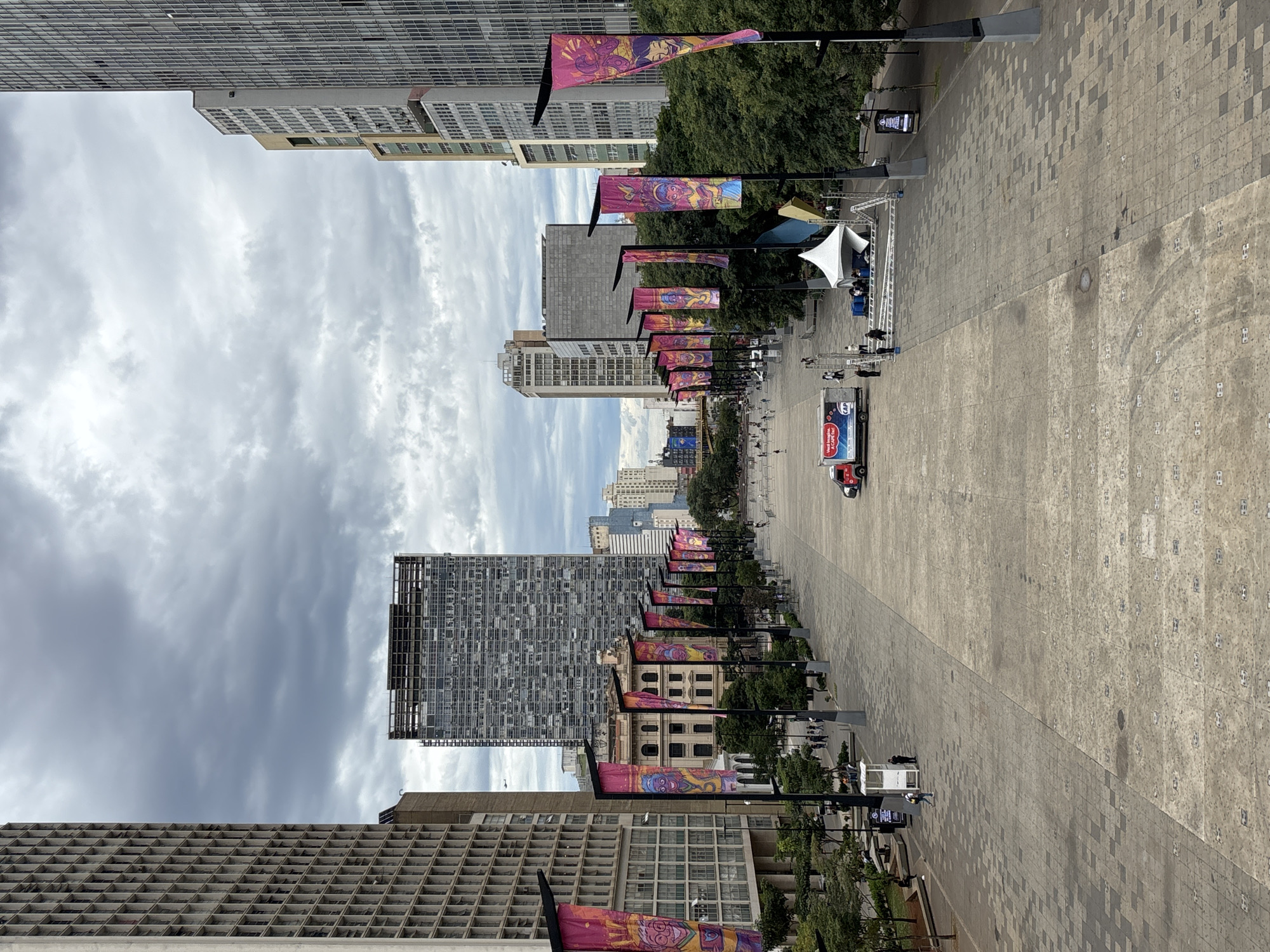 The image captures a wide pedestrian walkway in São Paulo, Brazil, lined with lamp posts featuring colorful banners. High-rise buildings flank both sides of the walkway, some under construction, and a cloudy sky is visible above. The ground is paved with patterned stone, leading to a stage structure further down the path, and there's a truck with advertising visible. People are sparsely distributed along the walkway, indicating limited activity.