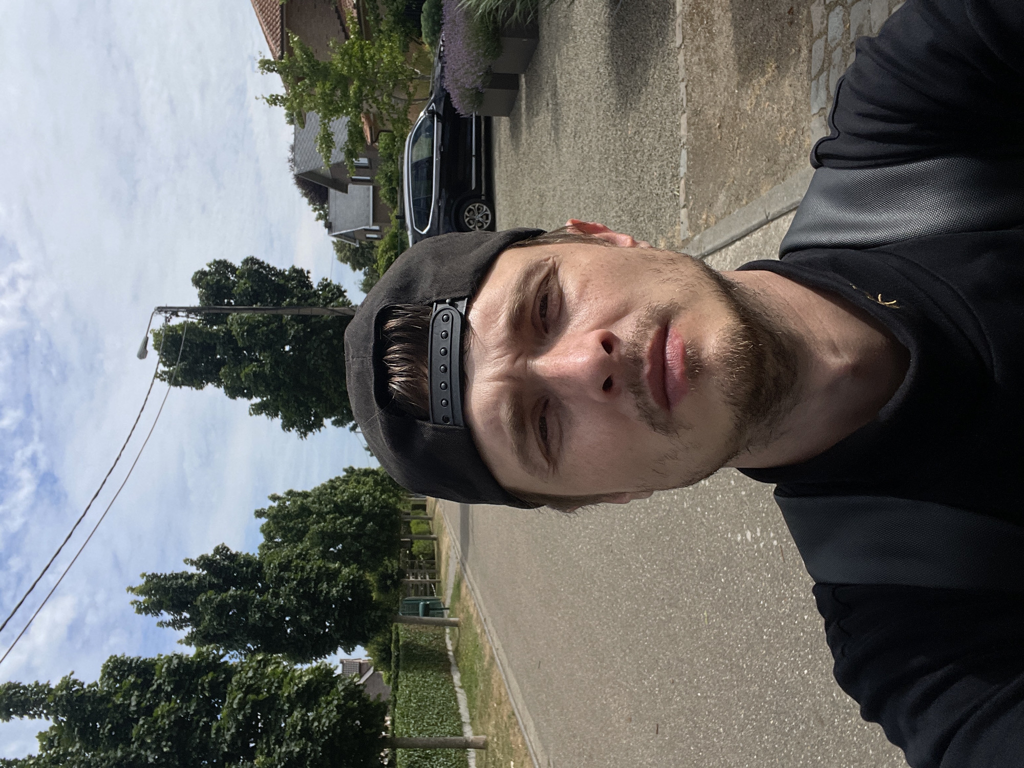 The image is a selfie taken outdoors on a street in Diepenbeek, Belgium. The subject, a man wearing a black baseball cap and a black shirt, is positioned in the foreground, facing the camera. Behind him, a paved road runs parallel to a neatly trimmed green hedge on the left side and a gravel driveway with lavender planters and houses on the right. A dark colored SUV is parked on the driveway. The sky is partly cloudy, and power lines are visible overhead, stretching across the frame. The scene suggests a residential area with well-maintained properties and a calm, daytime atmosphere.