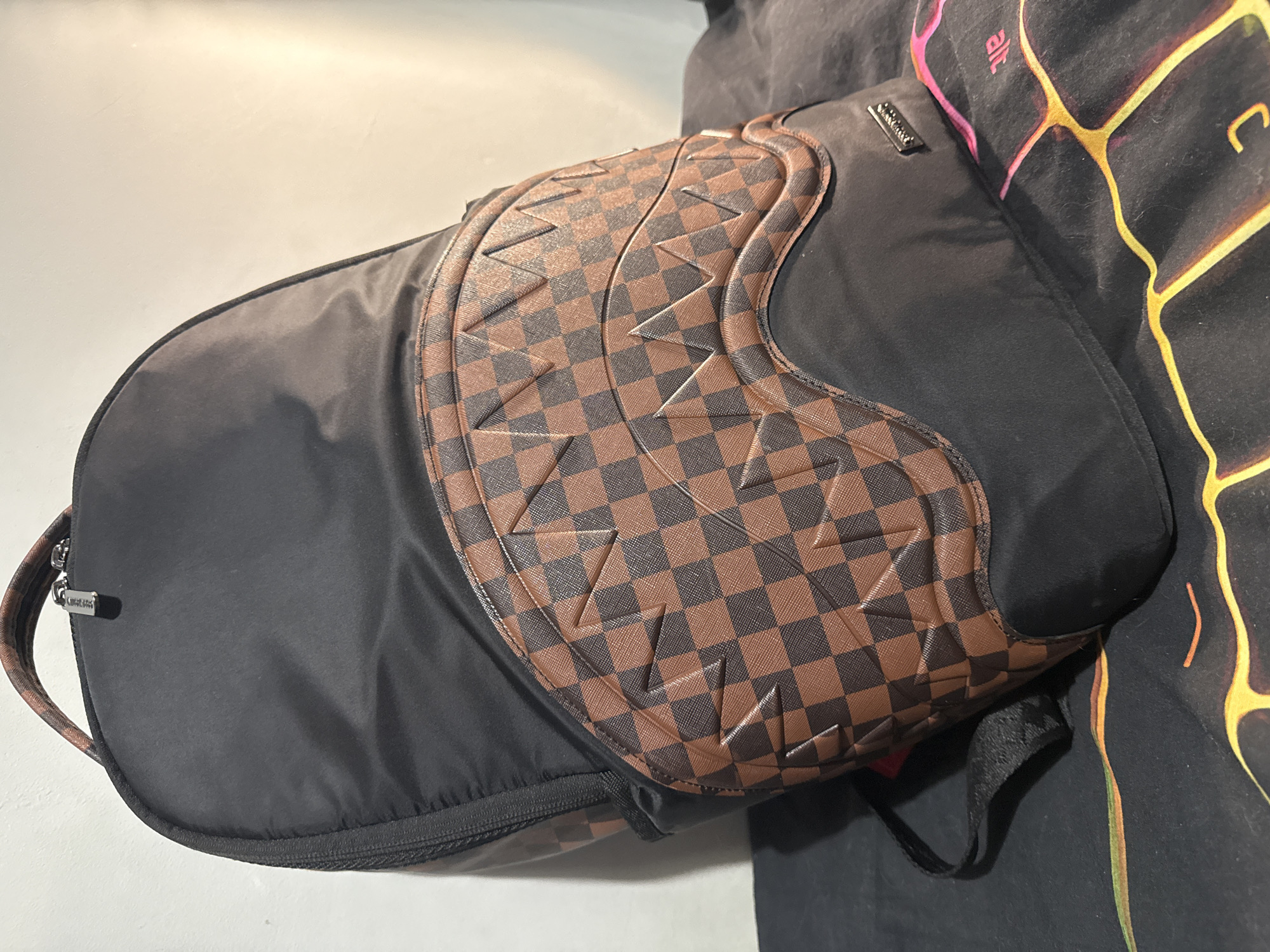 Here is a detailed description of the image:
The image shows a black backpack with brown checkered accents, partially open and leaning against a dark, patterned garment. The backpack's main body is made of black fabric, with a curved flap featuring a brown checkered pattern reminiscent of a racing design. The flap is decorated with raised, triangular details. The backpack has a zipper closure on its left side, and a small metal brand plate is visible near the top. The backpack appears to be placed near a surface with a black cloth with pink and yellow designs. The backdrop is a plain, light-colored wall.