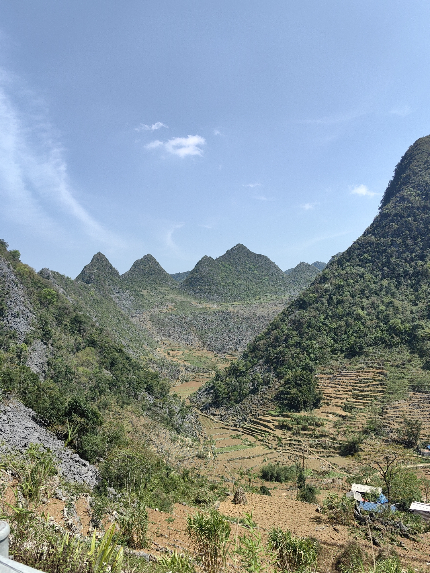 The image depicts a mountainous landscape in Lũng Cú Commune, Vietnam. The scene features a valley surrounded by verdant, rugged hills and mountains, some with visible rocky outcrops. Terraced fields are carved into the slopes, and a few buildings are situated in the lower right corner. The sky is blue with some scattered clouds.